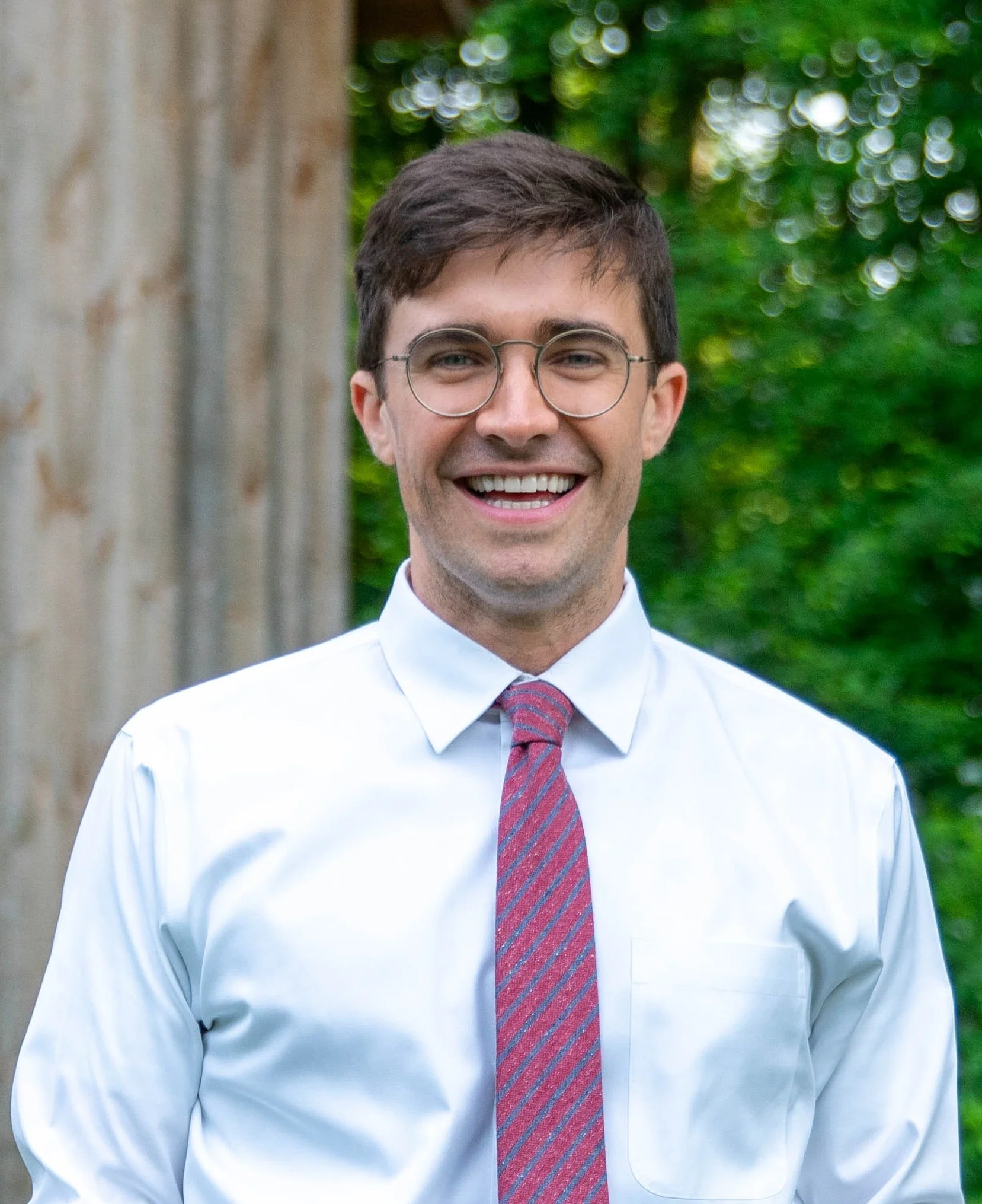 A smiling young man wearing glasses, a white dress shirt, and a striped tie outdoors with a background of blurred green trees and a wooden structure.