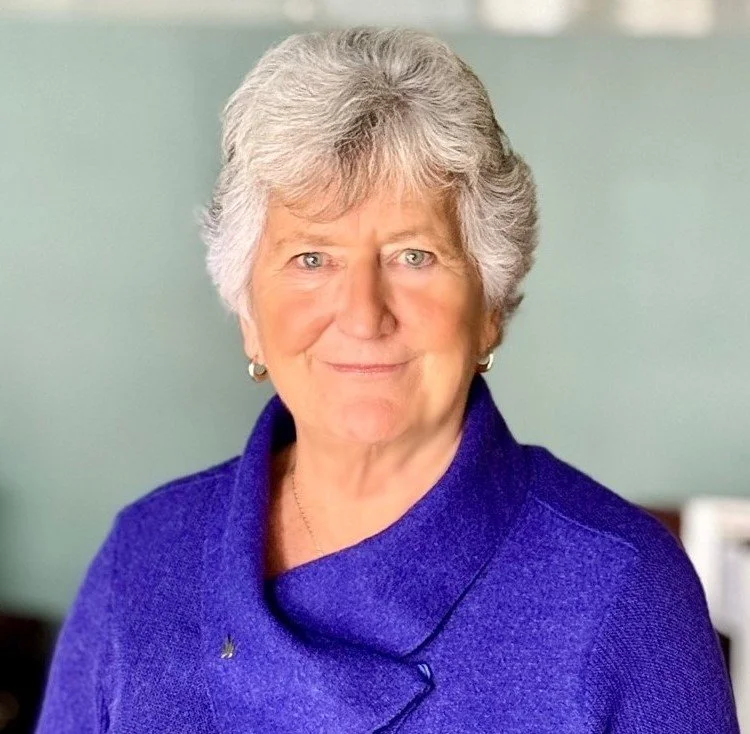 Close-up of an older woman with short gray hair, wearing a royal blue jacket, smiling softly in a well-lit indoor setting.
