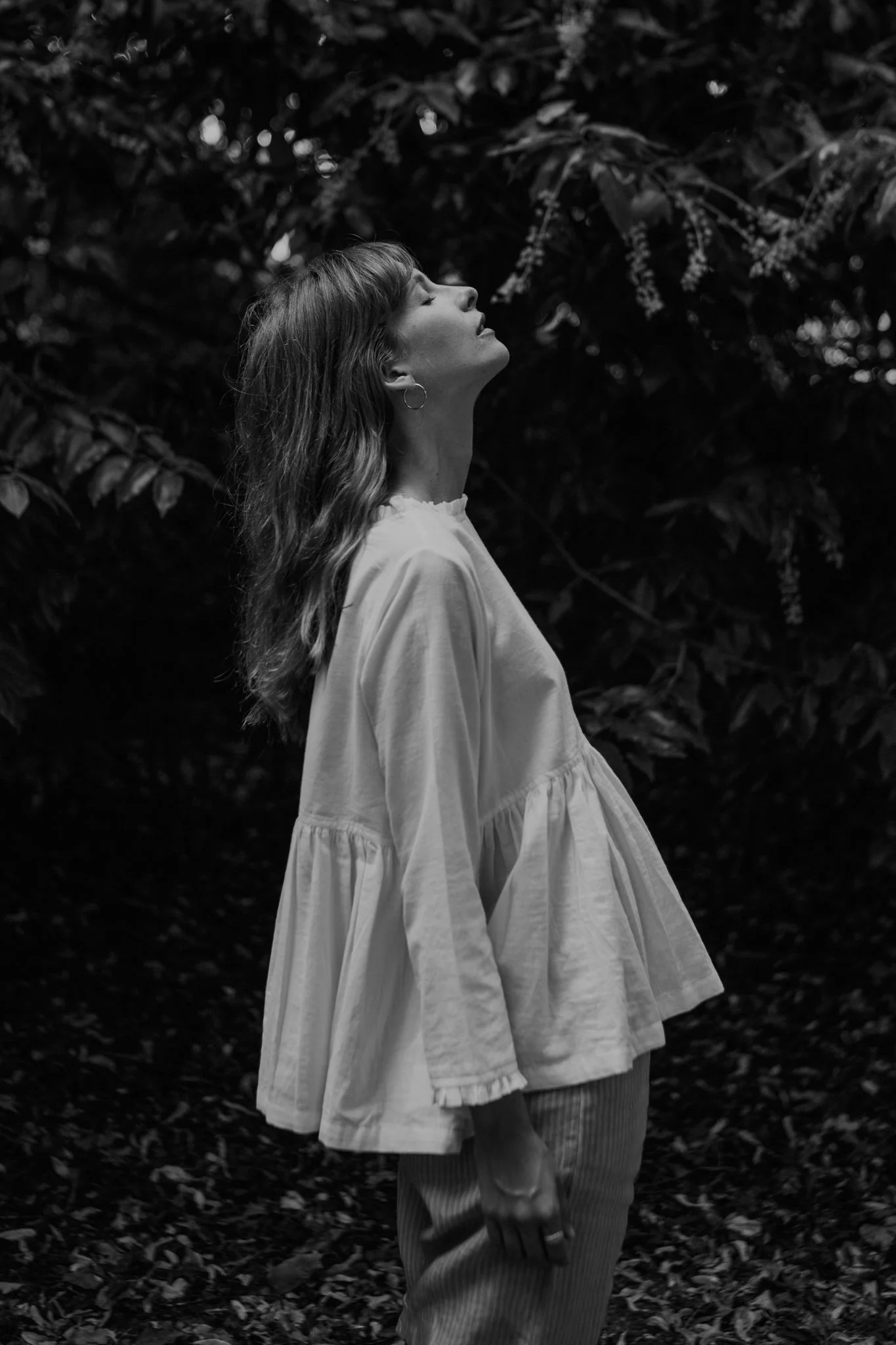 Black and white photo of a woman standing with her eyes closed, head tilted back, surrounded by foliage.