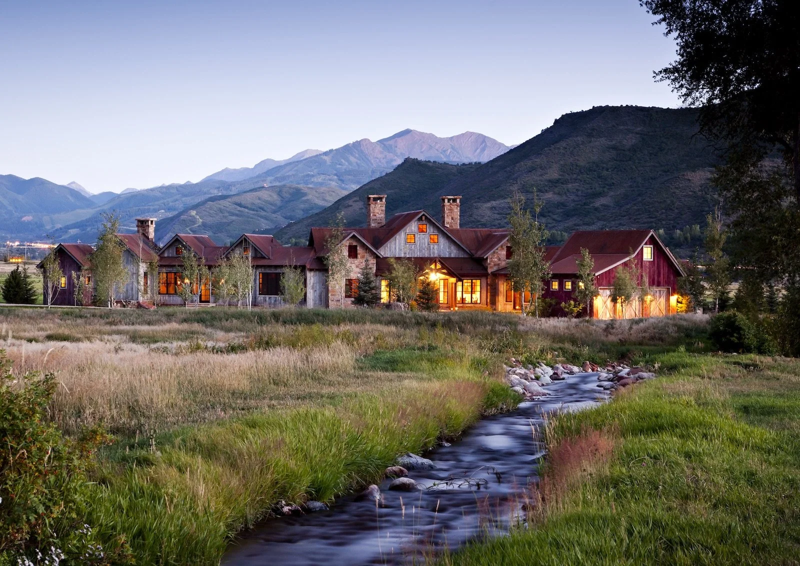 A rustic mountain house with warm interior lights, situated in a lush green meadow with a small creek in the foreground, surrounded by trees and mountains in the background at dusk.