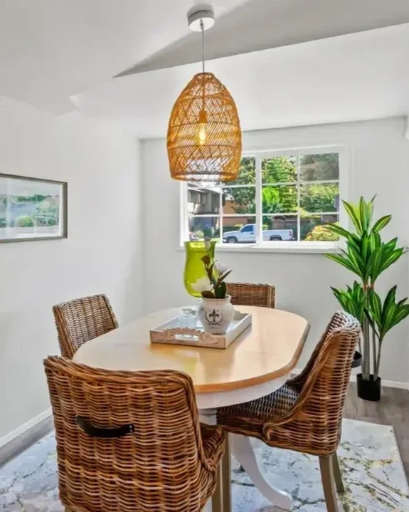 A cozy dining room with a white oval table surrounded by four wicker chairs, a wicker pendant light hanging from the ceiling, a potted plant and decorative vase on the table, a large window with a view of a residential street, and a framed landscape picture on the wall.