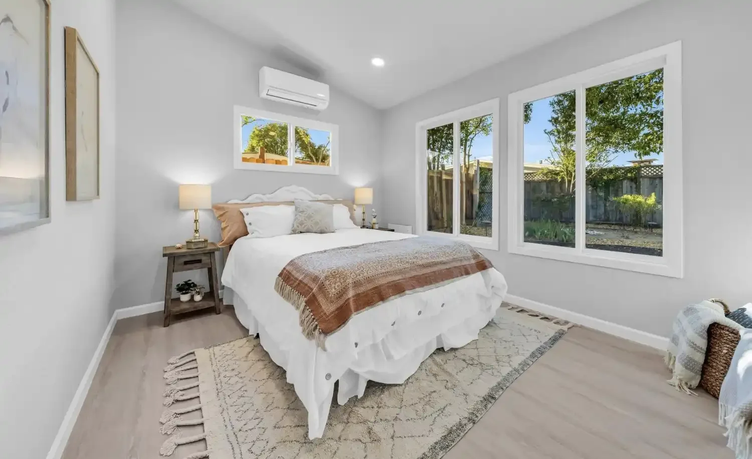 A bedroom with a white bed, beige and white bedding, two nightstands with lamps, large windows showing a backyard, and light wood flooring.