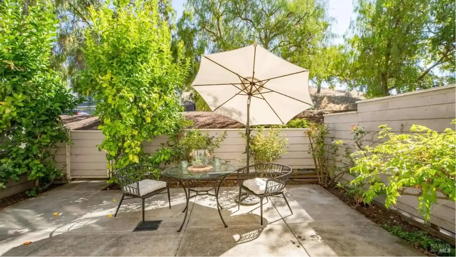 Small backyard patio with a round glass table, two black metal chairs with cushions, and a large white patio umbrella. There are plants and trees surrounding the patio, with a wooden fence and hilly landscape in the background.