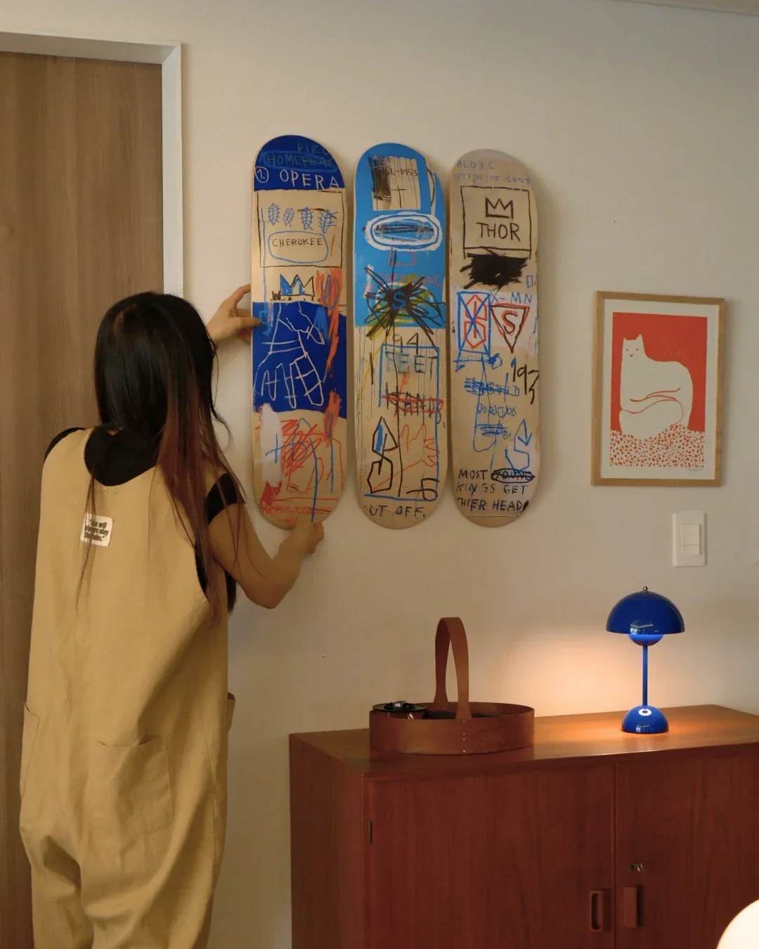 A woman with long hair, wearing a beige apron over a black shirt, is hanging artwork on a white wall. The artwork includes three tall, skateboard-shaped pieces with colorful, childlike drawings and writings. To the right, there is a framed picture of a white cat on a red background, and below that, a small blue desk lamp on a wooden cabinet.