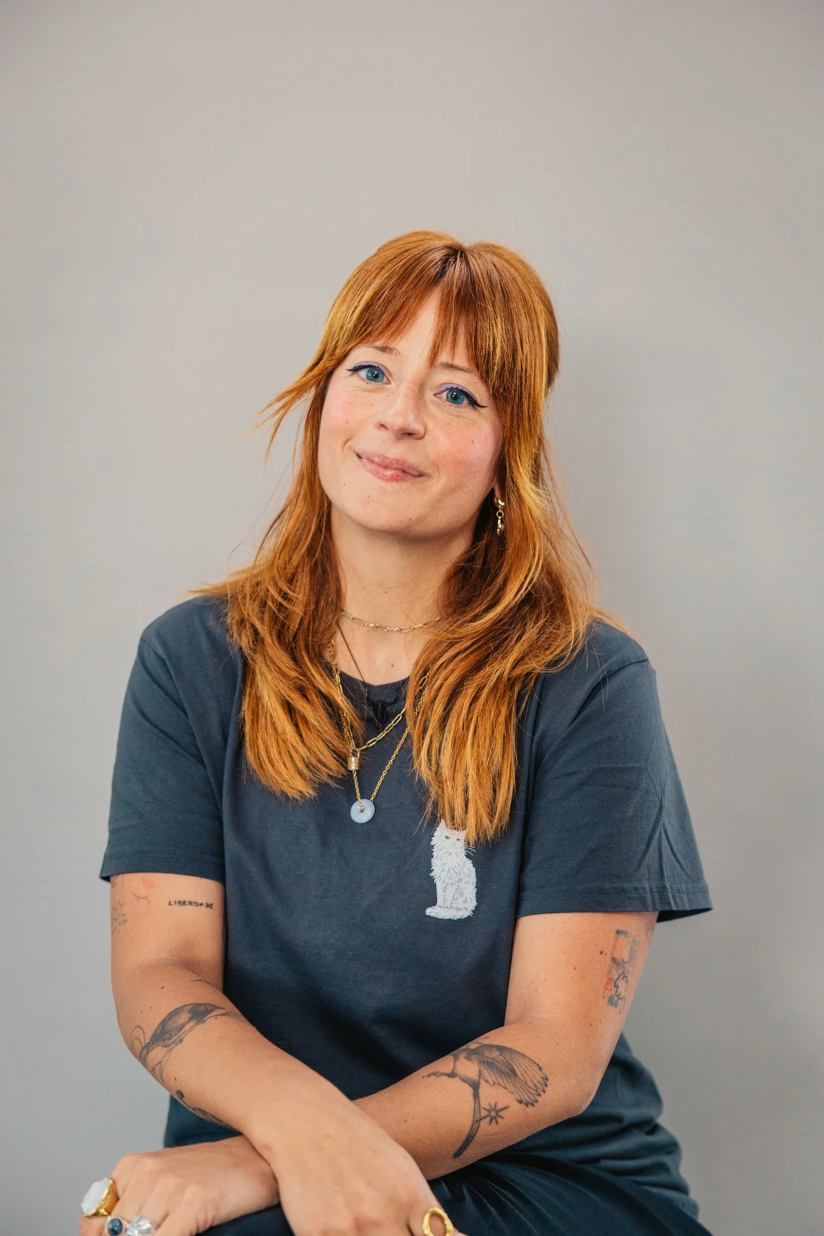 A woman with red hair and blue eyes, smiling, wearing a dark t-shirt with a white dog silhouette, sitting against a plain light gray background.