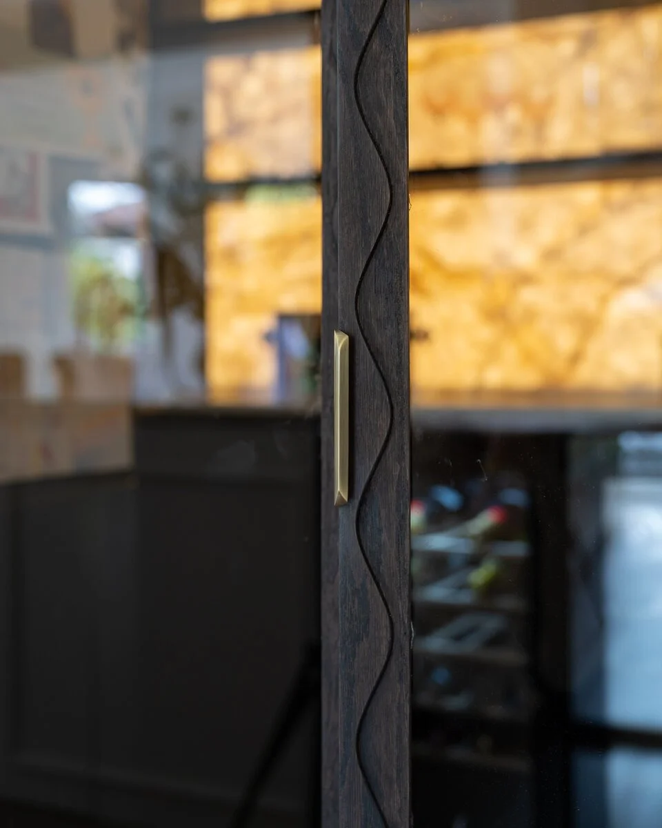 Brass handle detail on dark oak cabinetry with an illuminated drinks cabinet beyond.