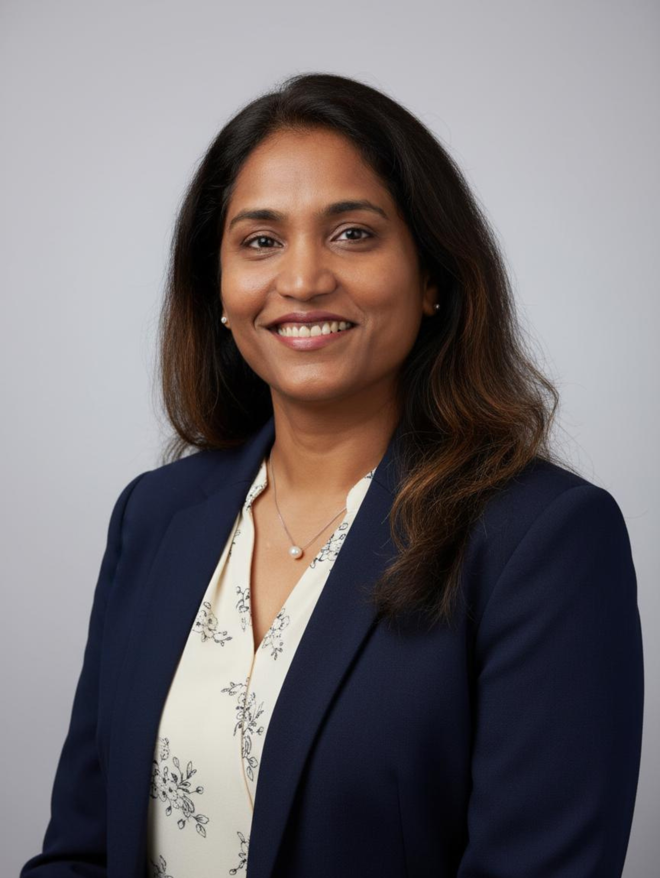A professional woman with long dark hair smiling, wearing a navy blazer, a cream blouse with floral pattern, pearl earrings, and a pearl necklace, against a plain gray background.