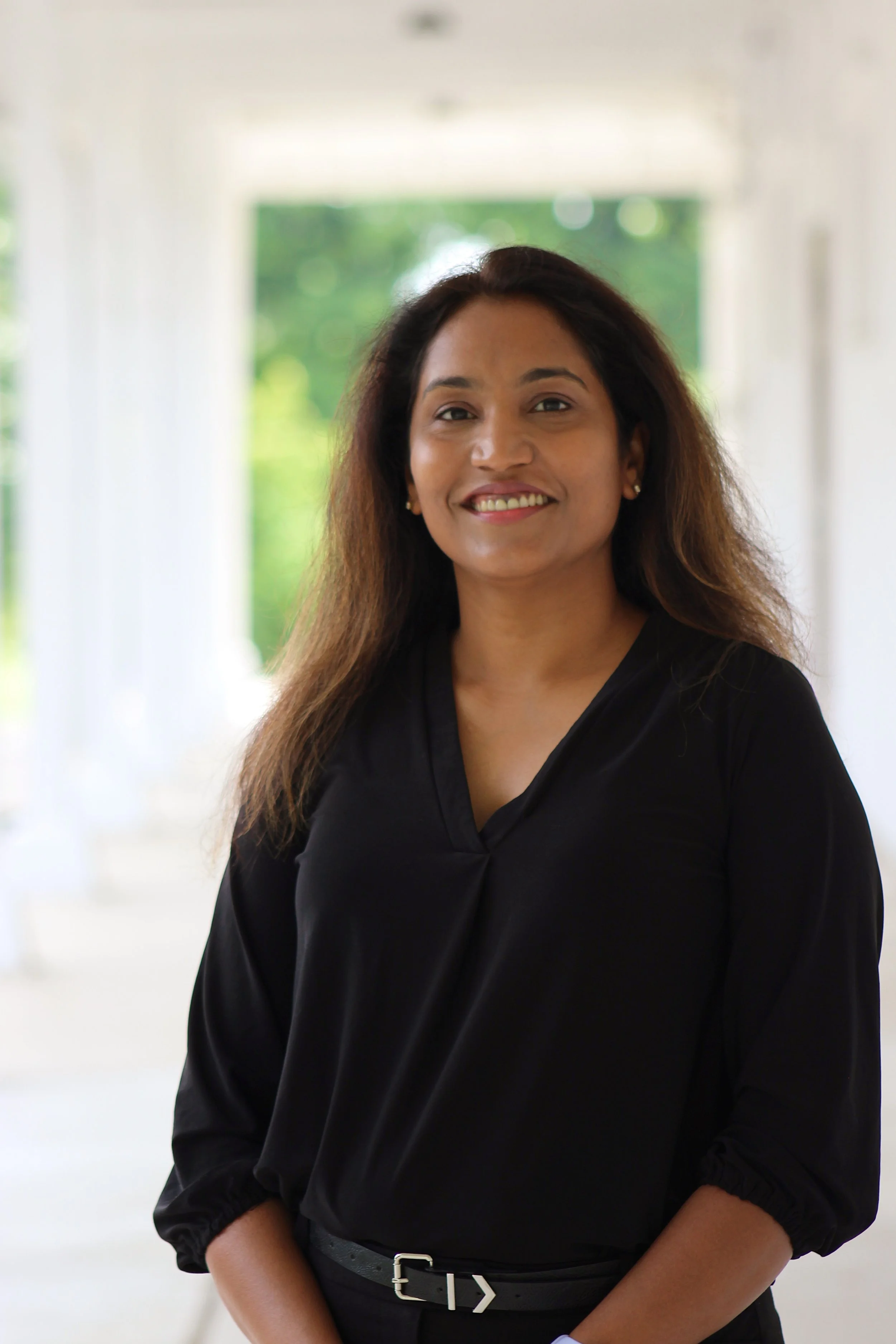 A woman with long hair wearing a black blouse and belt, smiling, standing in a bright, white corridor with windows showing green trees outside.