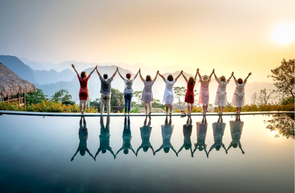 Group of nine friends standing on a bridge holding hands at sunset, with their reflection visible in a pool of water, mountains in the background.