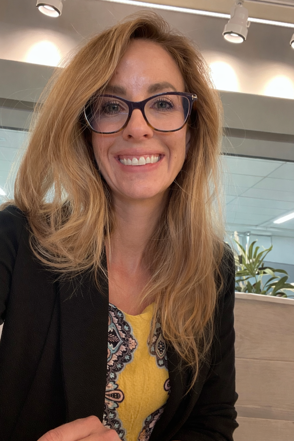 A smiling woman with glasses and long red hair, wearing a black blazer and a yellow dress with a paisley pattern, sitting indoors with a plant and modern ceiling lights in the background.