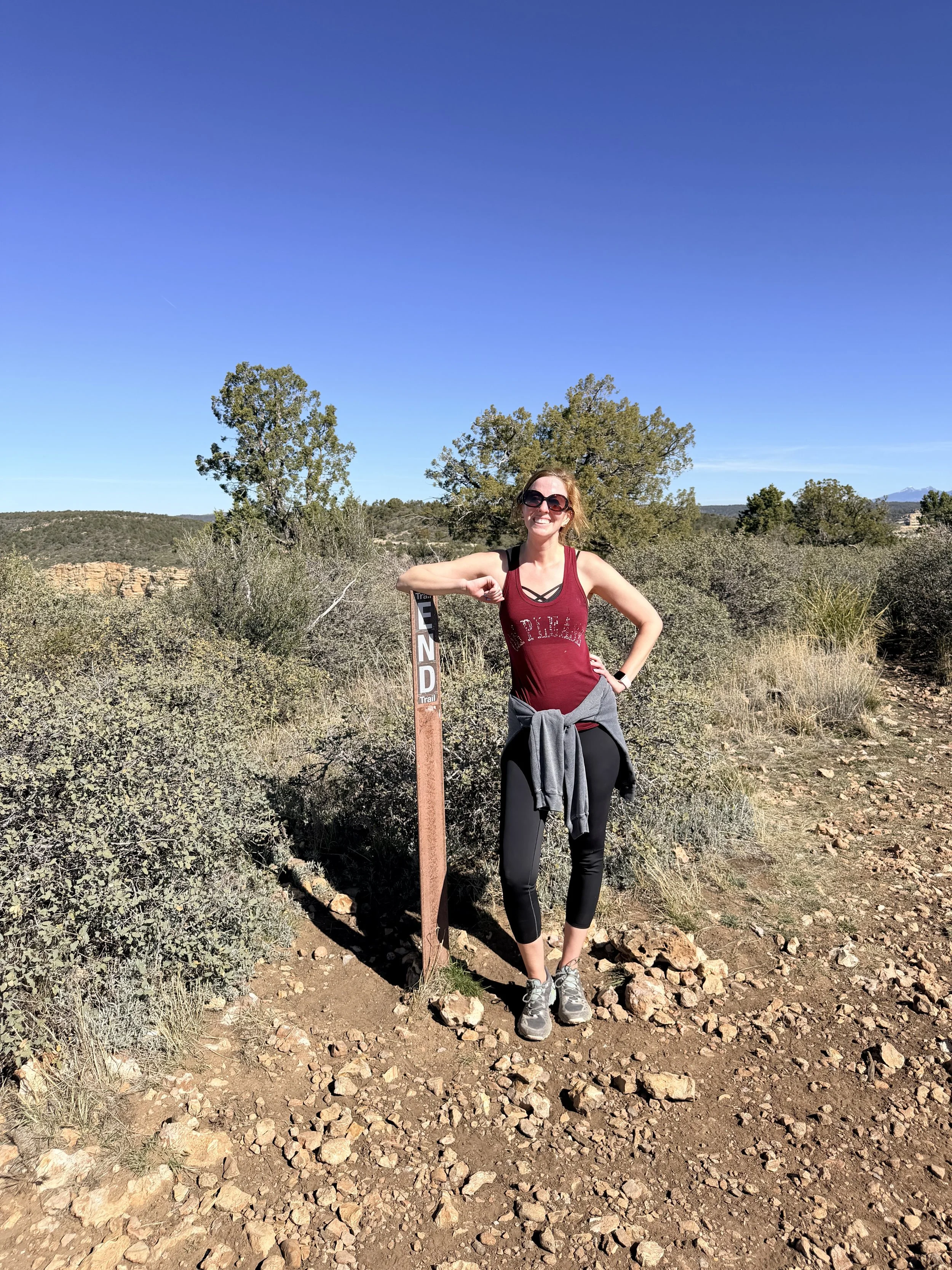 A woman standing outdoors on a hiking trail with a scenic background of trees, rocky terrain, and a clear blue sky, smiling.