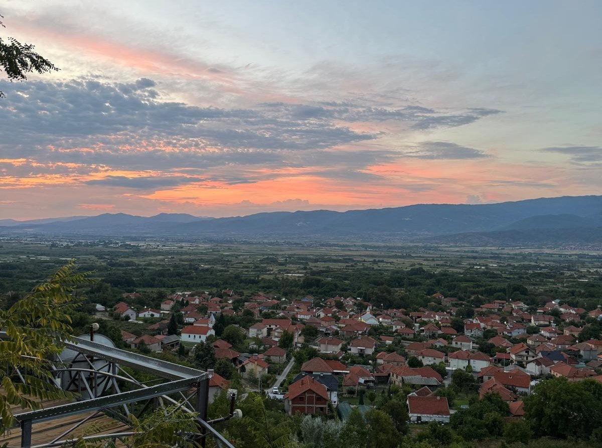 A scenic view of a suburban neighborhood with red-tiled roofs, green trees, and a mountain range in the distance under a colorful sky during sunset. North Macedonia, Strumica valley.