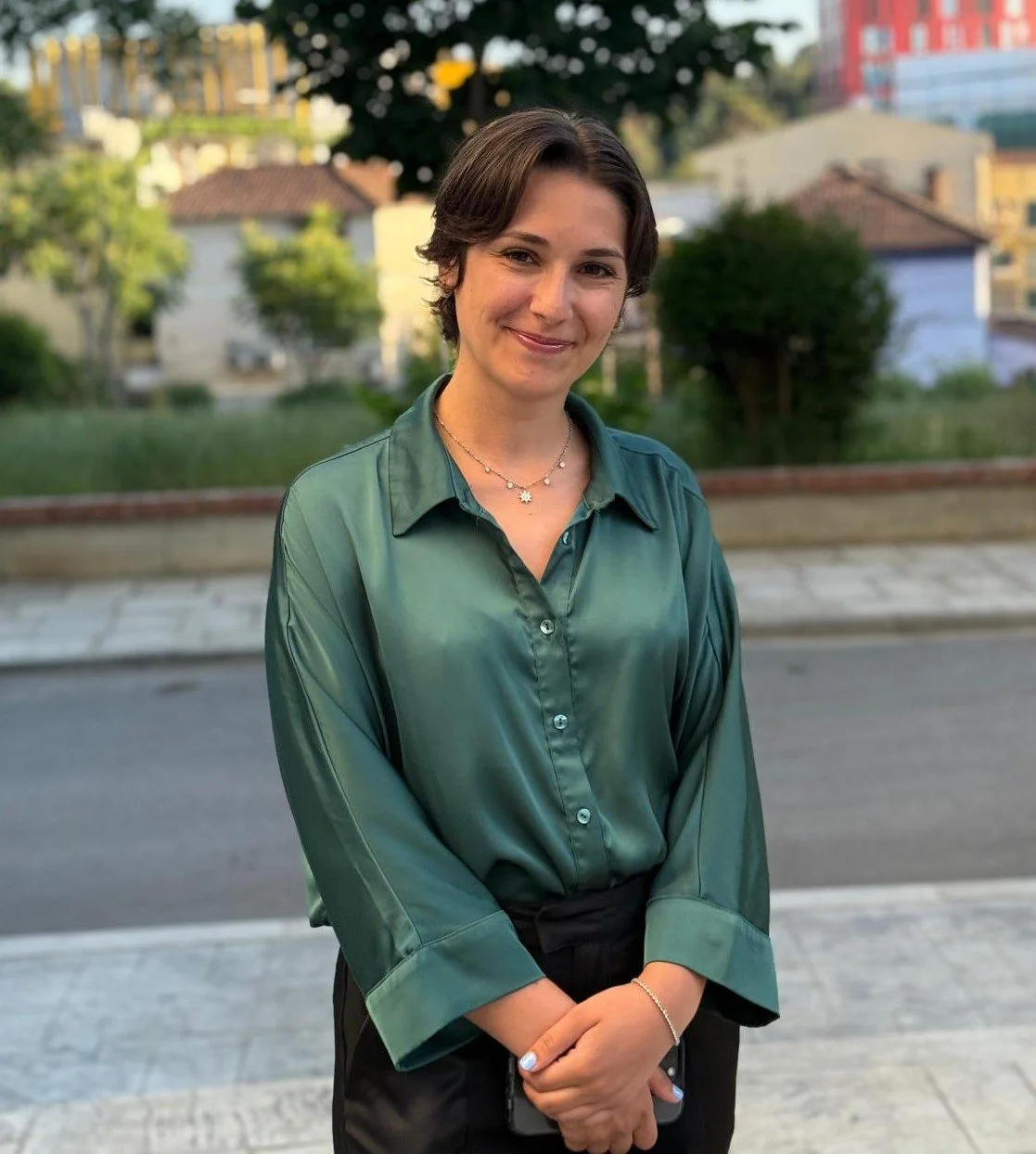 A woman with short dark hair, wearing a green satin blouse, standing outdoors in a neighborhood with trees and houses in the background, smiling.