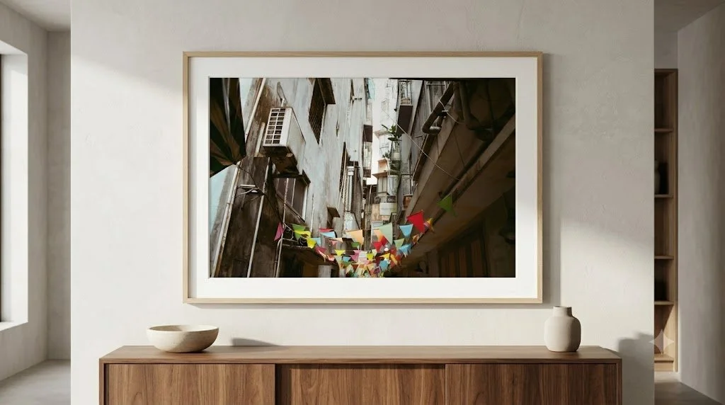 A framed photograph hanging on a white wall inside a room. The photo depicts a view of a narrow urban alleyway decorated with colorful triangular flags strung across between buildings.