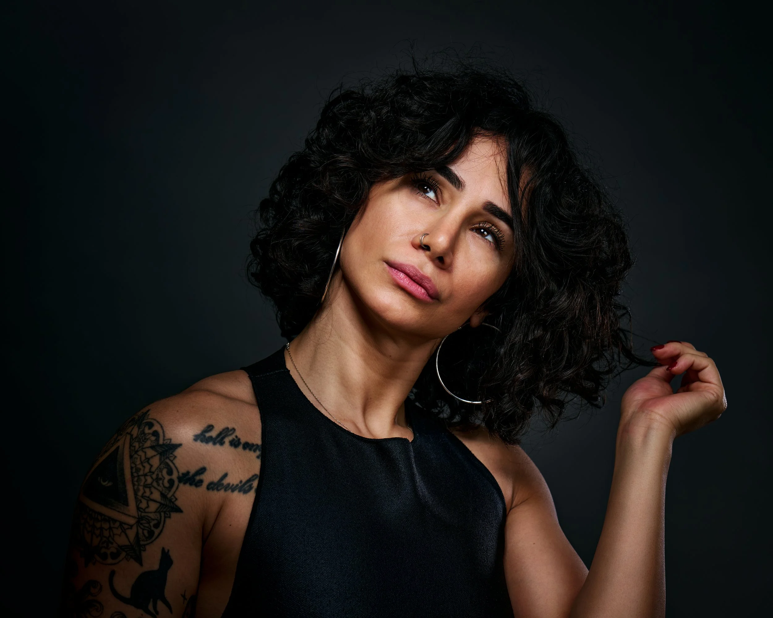 A woman with curly black hair, wearing large hoop earrings, a nose ring, and a sleeveless black top, looking slightly upward and to the side, with a tattoo on her left shoulder and upper arm, against a dark background.
