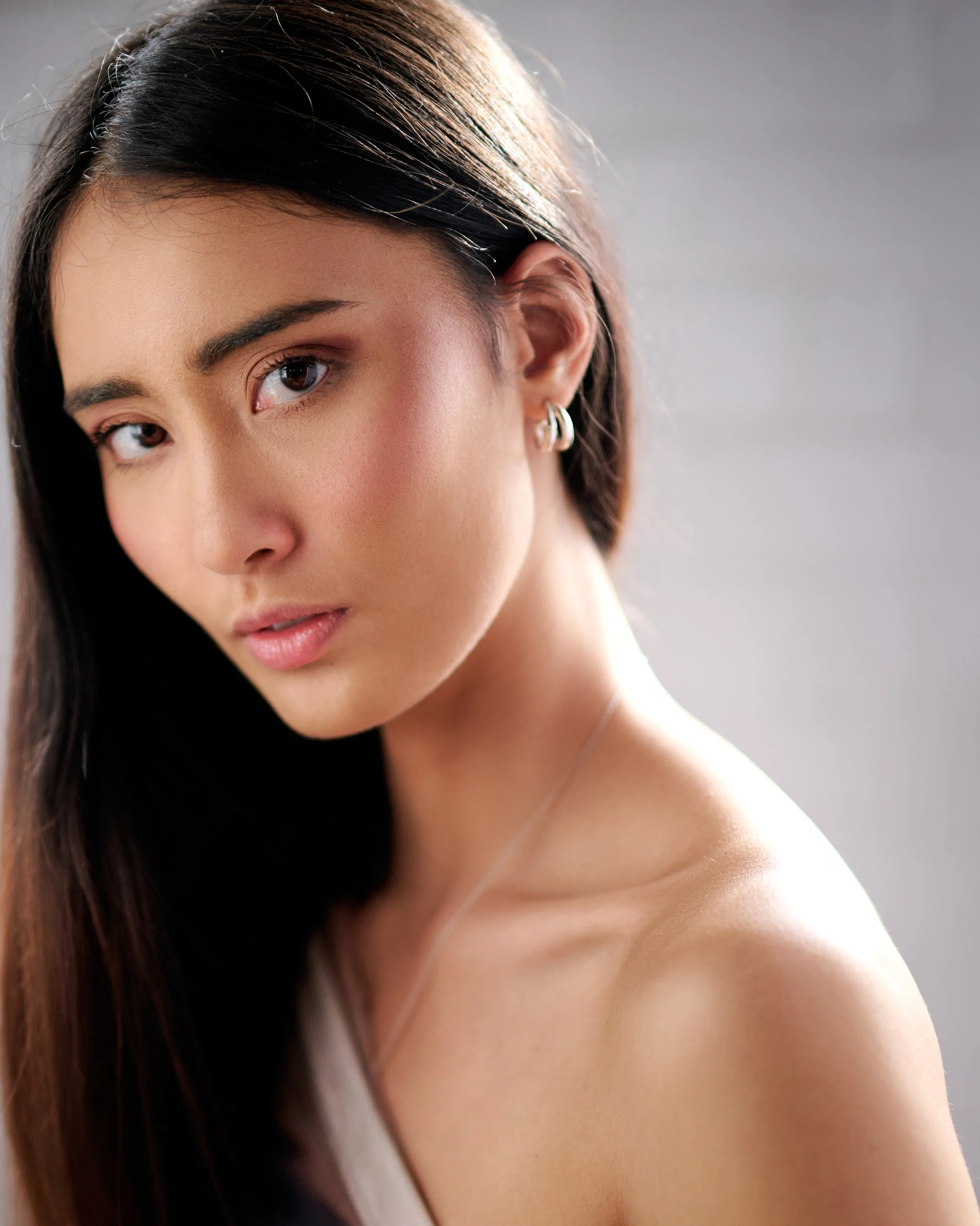 Close-up portrait of a young woman with long dark hair, wearing silver hoop earrings, looking into the camera with a soft expression.