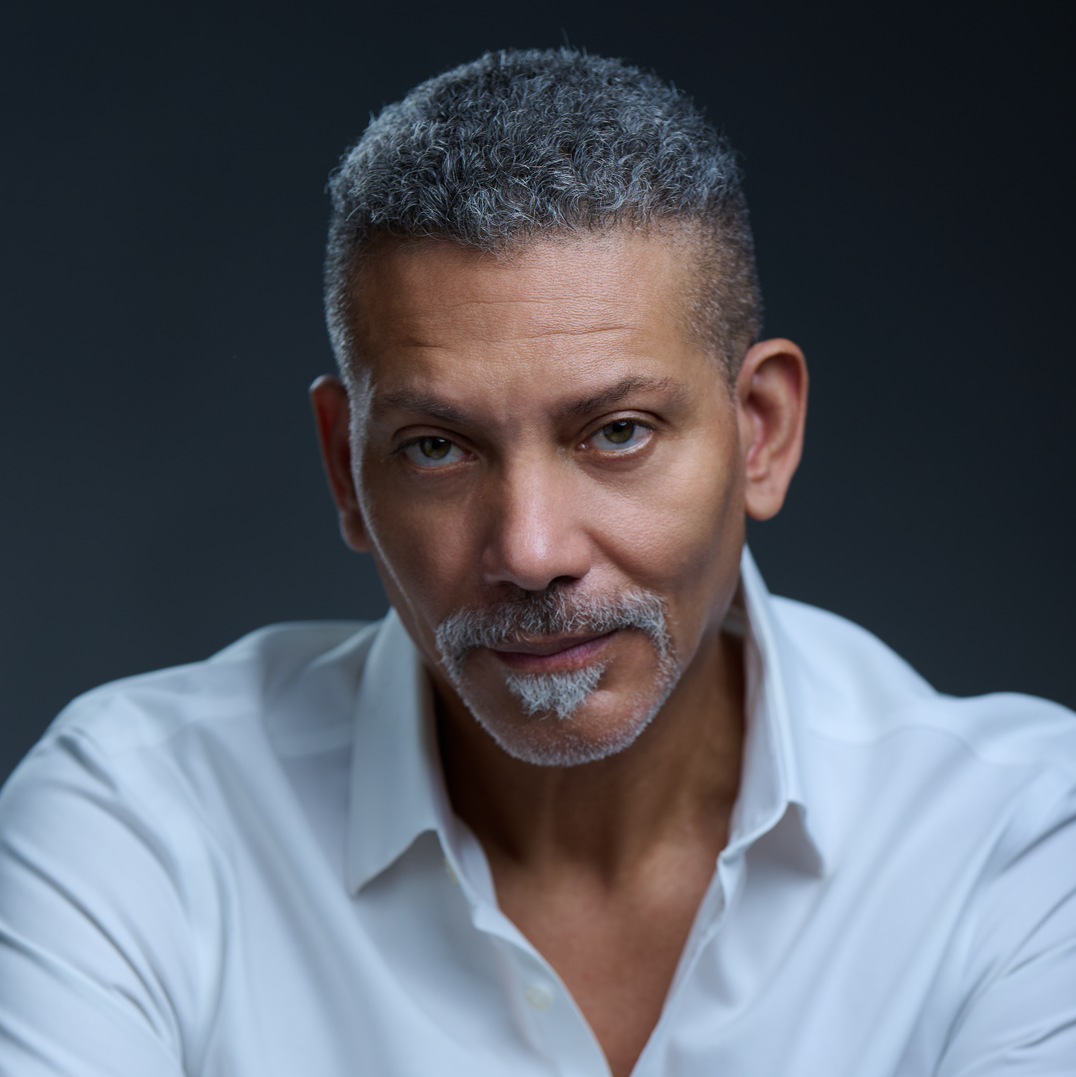 A middle-aged man with salt-and-pepper hair and beard, wearing a white shirt, looking directly at the camera against a dark background.