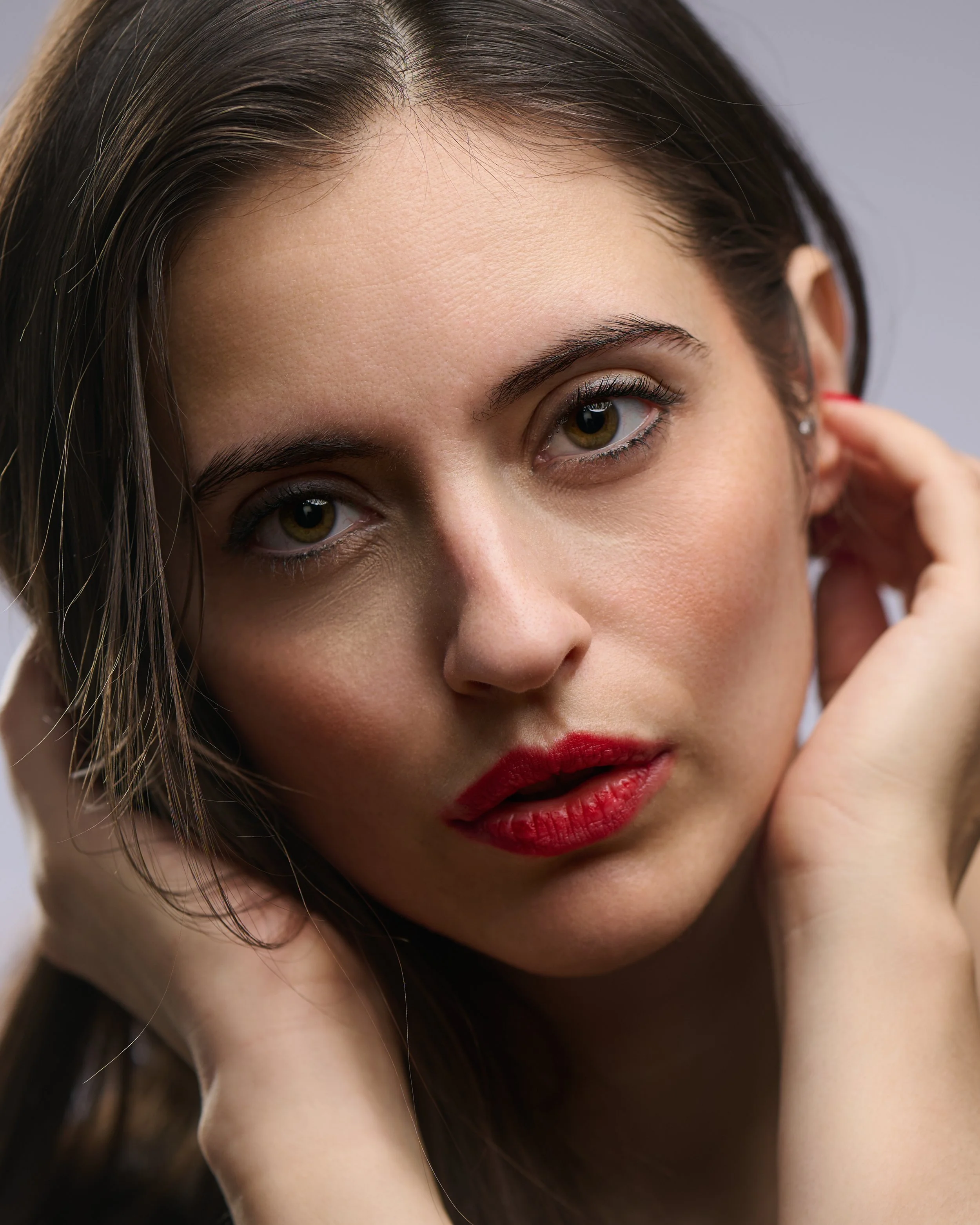 Close-up portrait of a woman with brown hair, wearing red lipstick, with one hand touching her face and the other near her ear, against a plain gray background.
