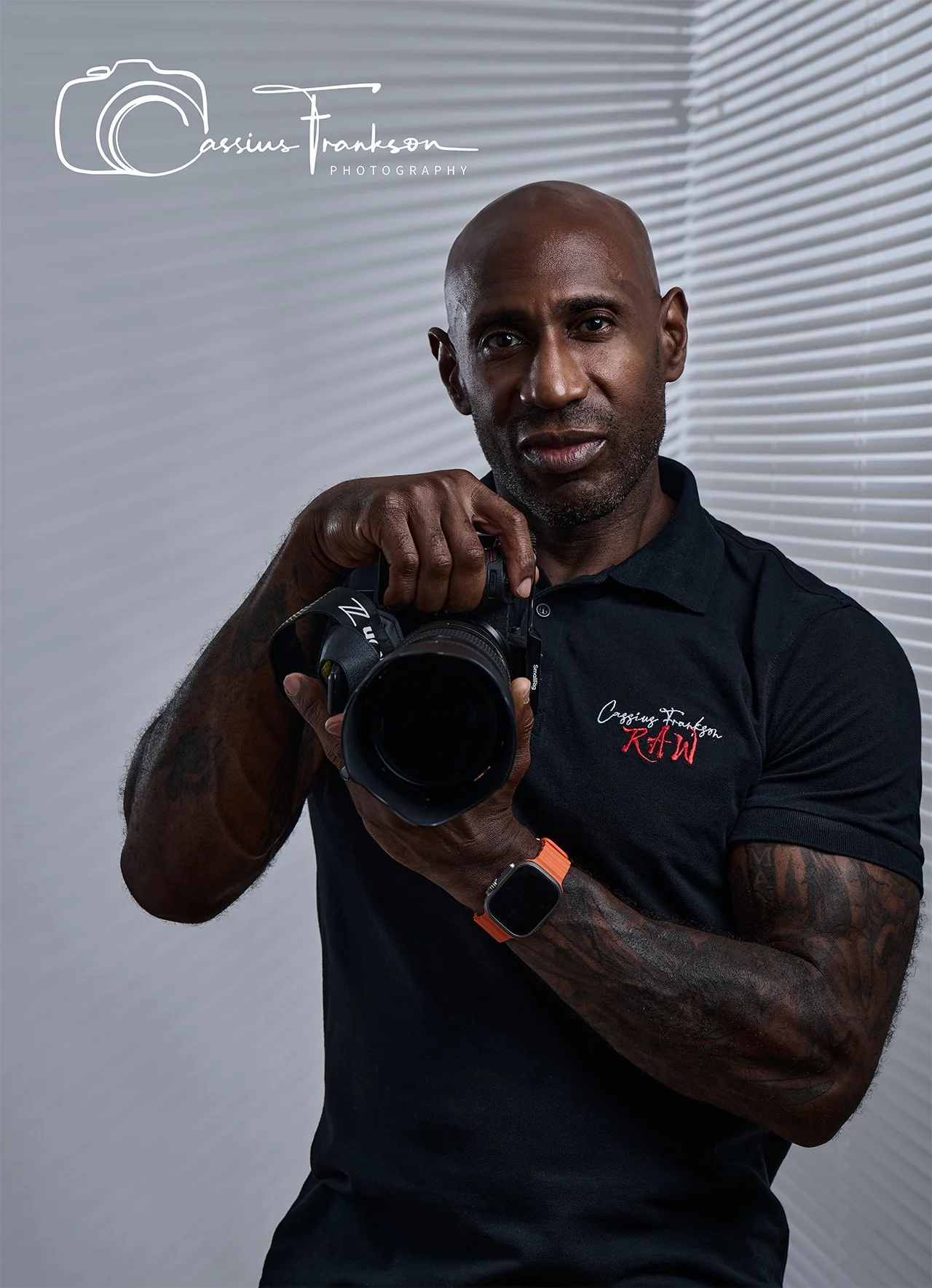 A man with tattoos on his arms holding a camera and taking a selfie. He is wearing a black shirt with red and white text and an orange smartwatch. The background features white blinds.