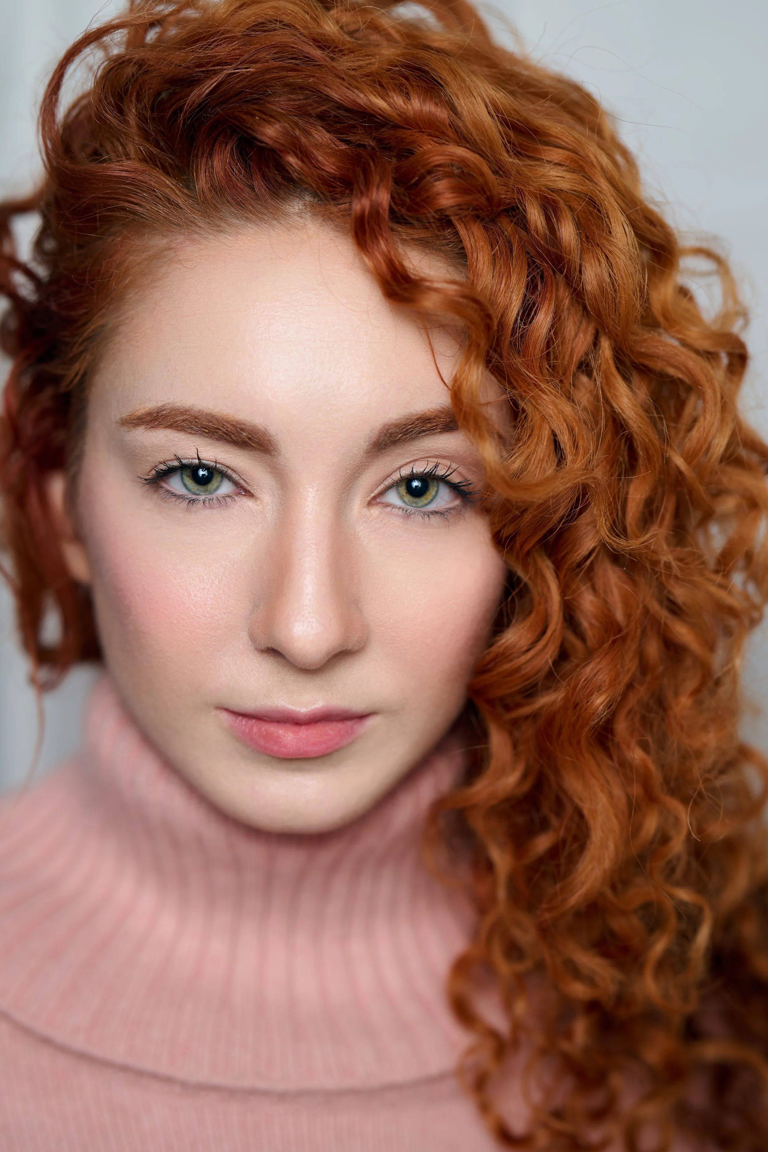 Close-up portrait of a woman with curly red hair, fair skin, and blue eyes wearing a pink turtleneck sweater.