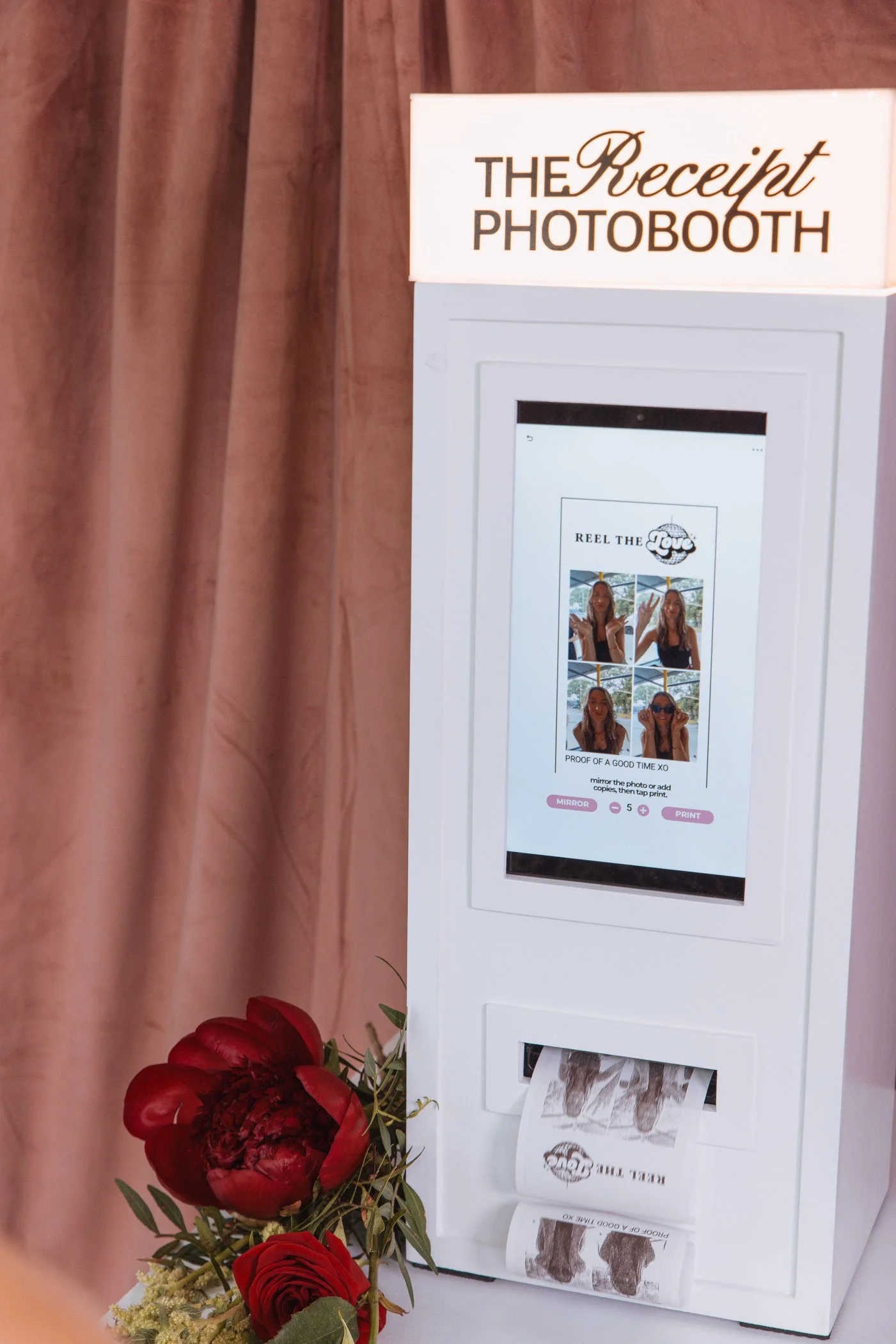 Photo of a photo booth with a sign that reads 'The Receipt Photobooth' and a screen showing a woman taking a picture, accompanied by a bouquet of red roses at the bottom left.