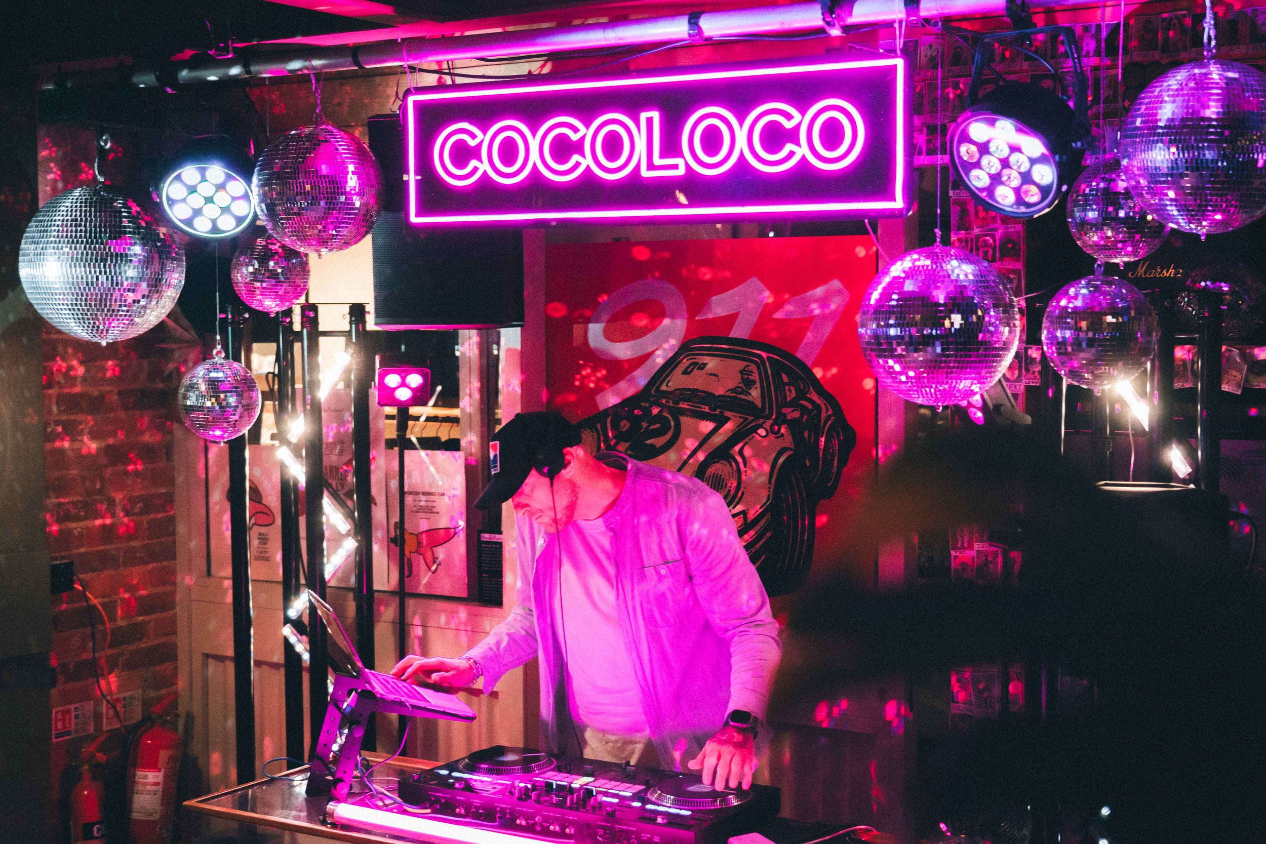 A DJ wearing a white jacket and black cap, using a laptop at a nightclub with pink neon lights, disco balls, and a red background with a car illustration, the word 'COCOLOCO' illuminated in pink neon above.