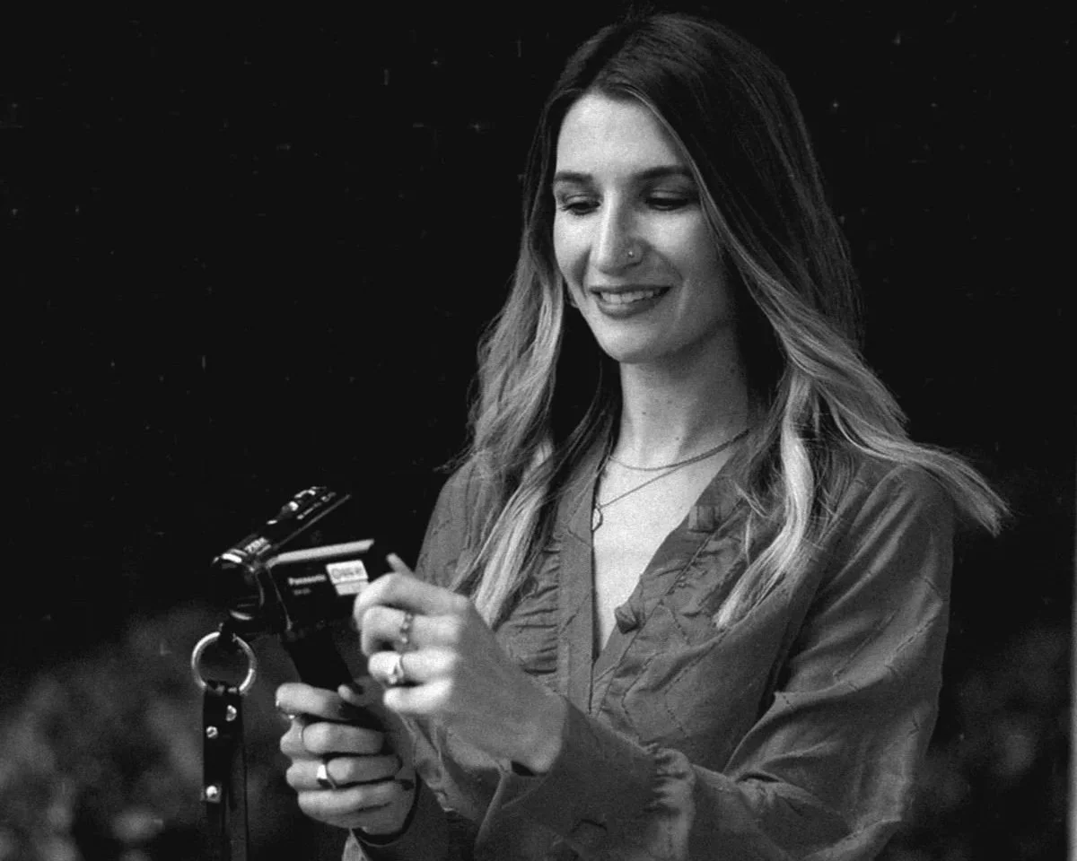 A woman with long wavy hair smiling while looking at a camera in her hands, against a dark background.