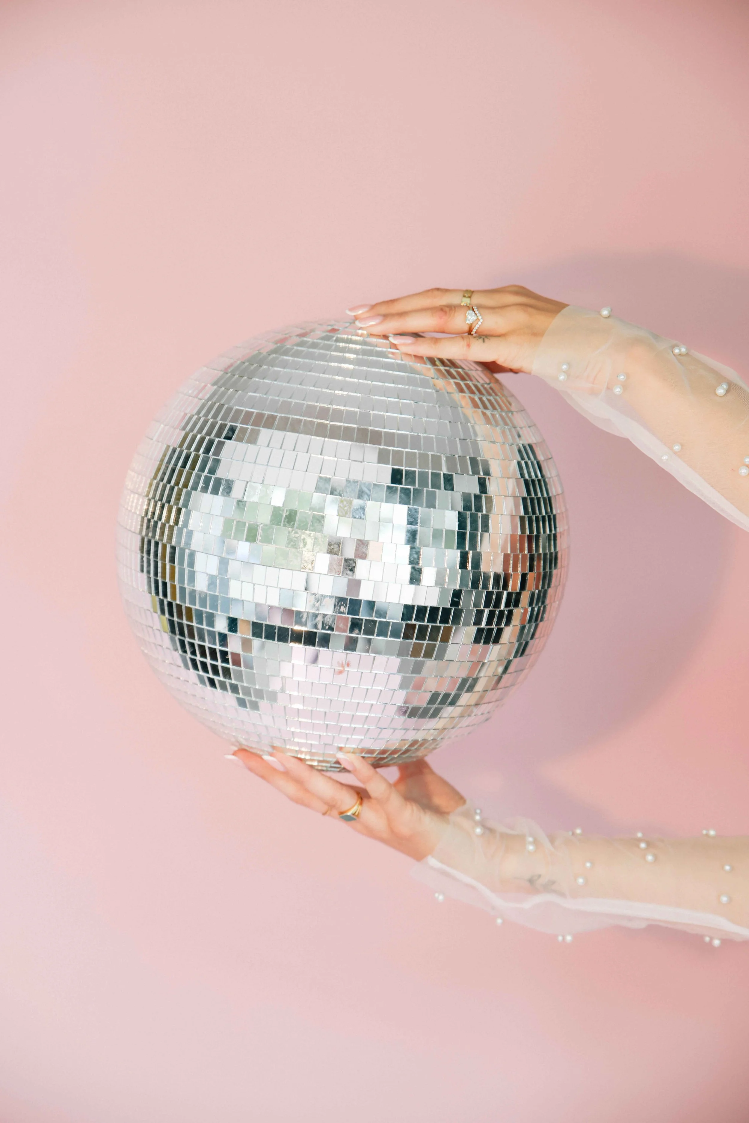 Person holding a large disco ball against a pink background, wearing a sheer long-sleeve top decorated with pearls.