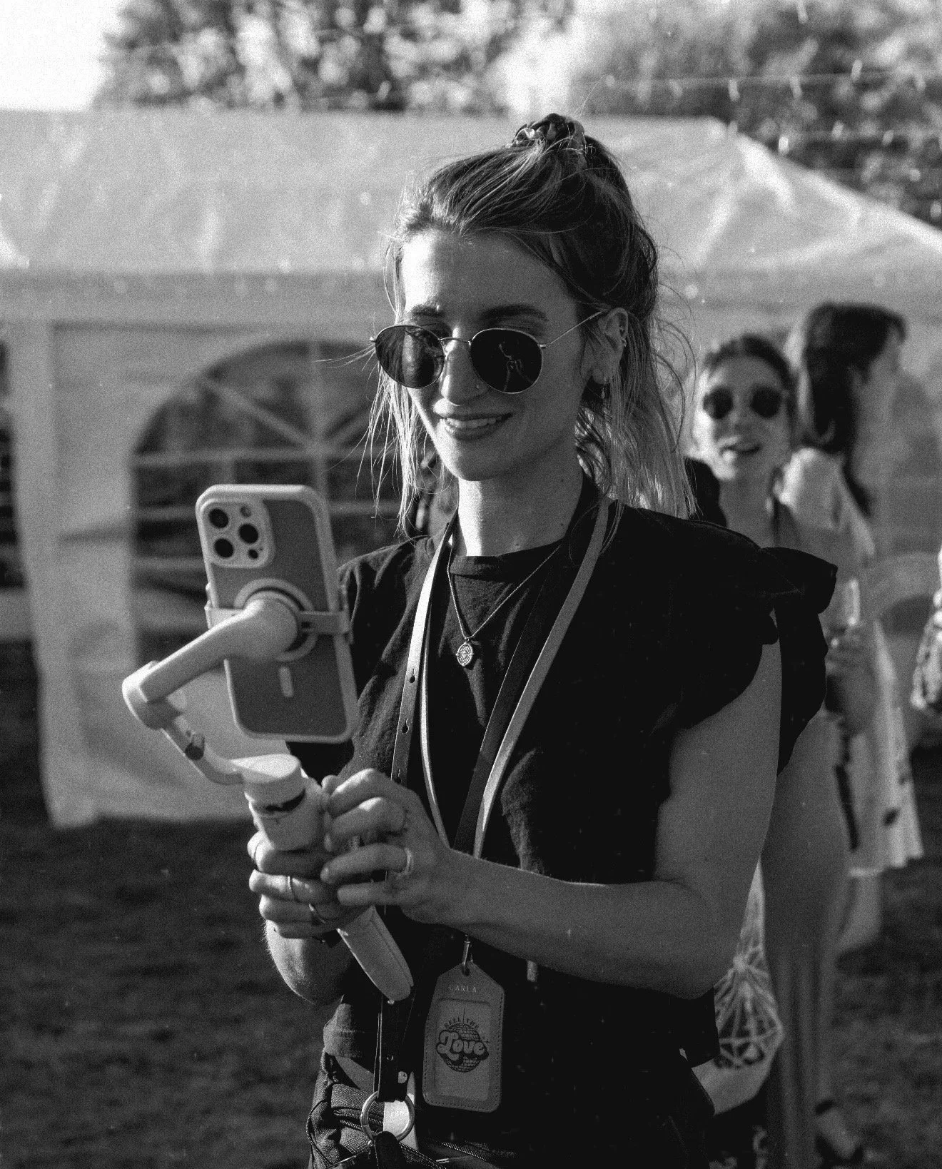 A woman with sunglasses, holding a smartphone mounted on a selfie stick, smiling, outdoors on a sunny day, with two other women in the background also wearing sunglasses and smiling.
