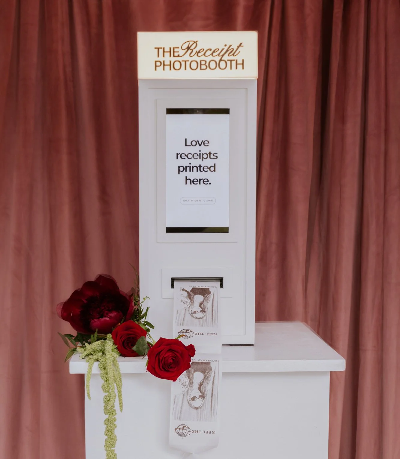 A photo booth titled 'The Receipt Photobooth' with a sign that says 'Love receipts printed here.' at a wedding or event. There are red roses and flowers at the base of the booth, with a pink curtain backdrop.