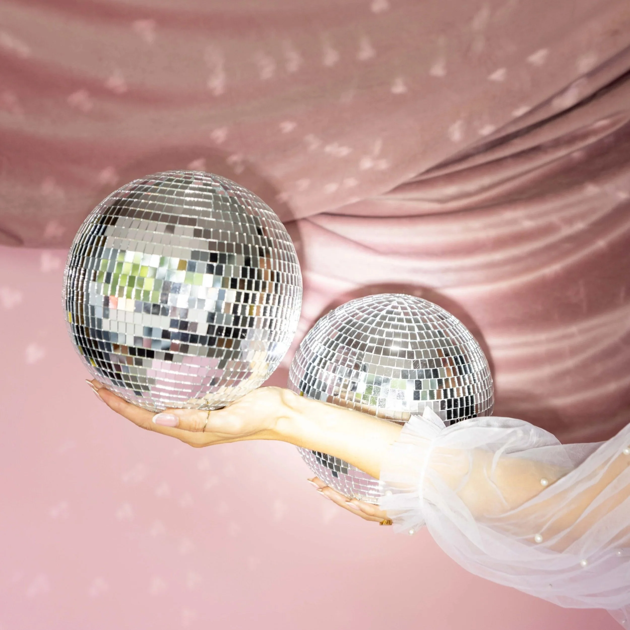 A hand holding three disco balls of different sizes against a pink, glittery backdrop.