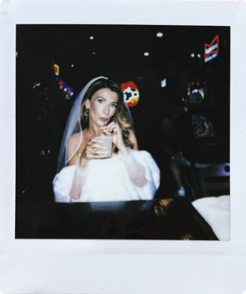 A woman dressed as a bride with a veil, holding a drink with a straw, in a dark setting with colorful, illuminated decorations in the background.