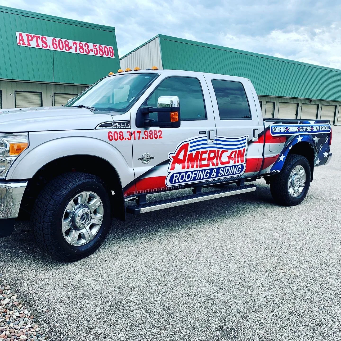 A white pickup truck with a patriotic patriotic-themed vinyl wrap advertising American Roofing & Siding. The wrap features red, white, and blue colors with stars and stripes. The truck displays contact information and services including roofing, sidi