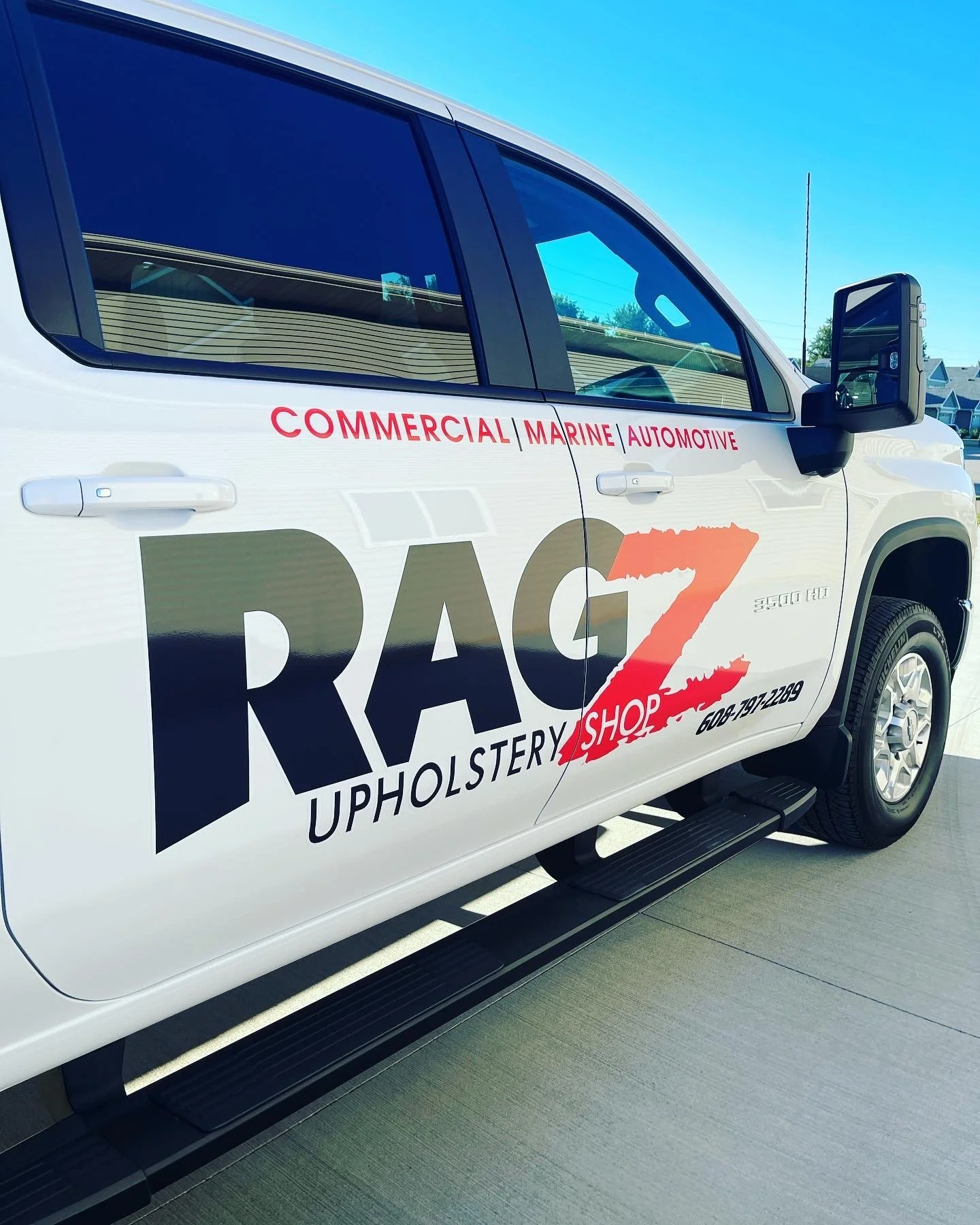 White truck with company branding for Rugz Upholstery Shop, including phone number, and services such as commercial, marine, and automotive upholstery, parked outside on a driveway.