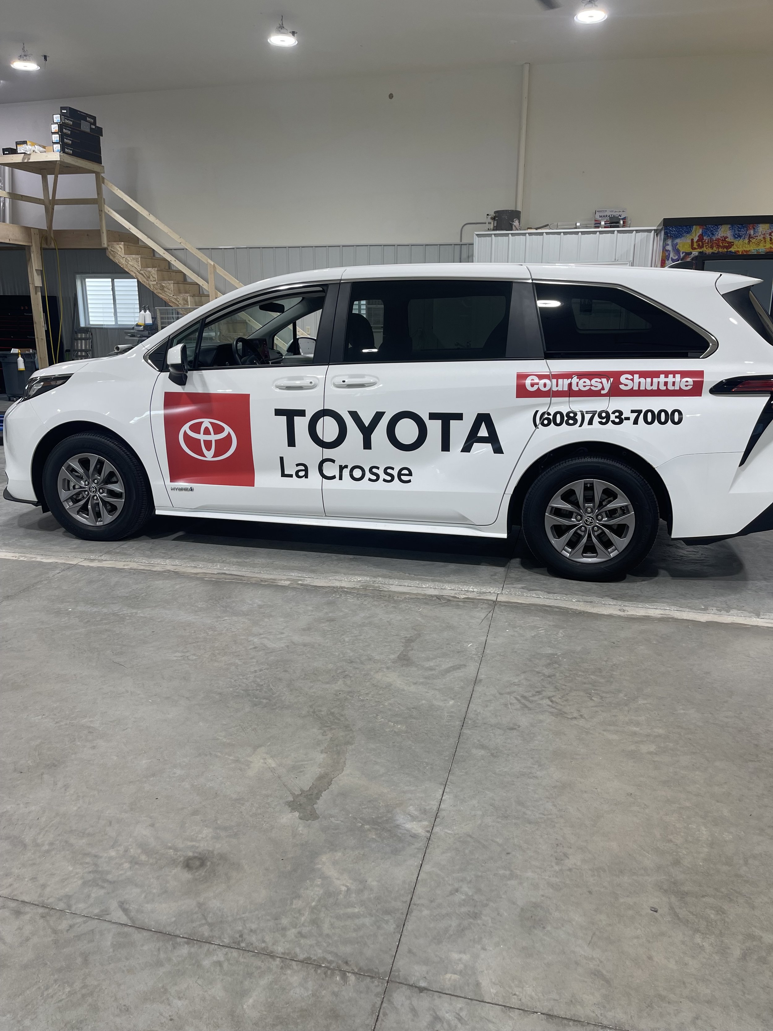 A white Toyota vehicle parked indoors with temporary advertising decals for Toyota La Crosse and courtesy shuttle, including a red Toyota logo, black text, and phone number.