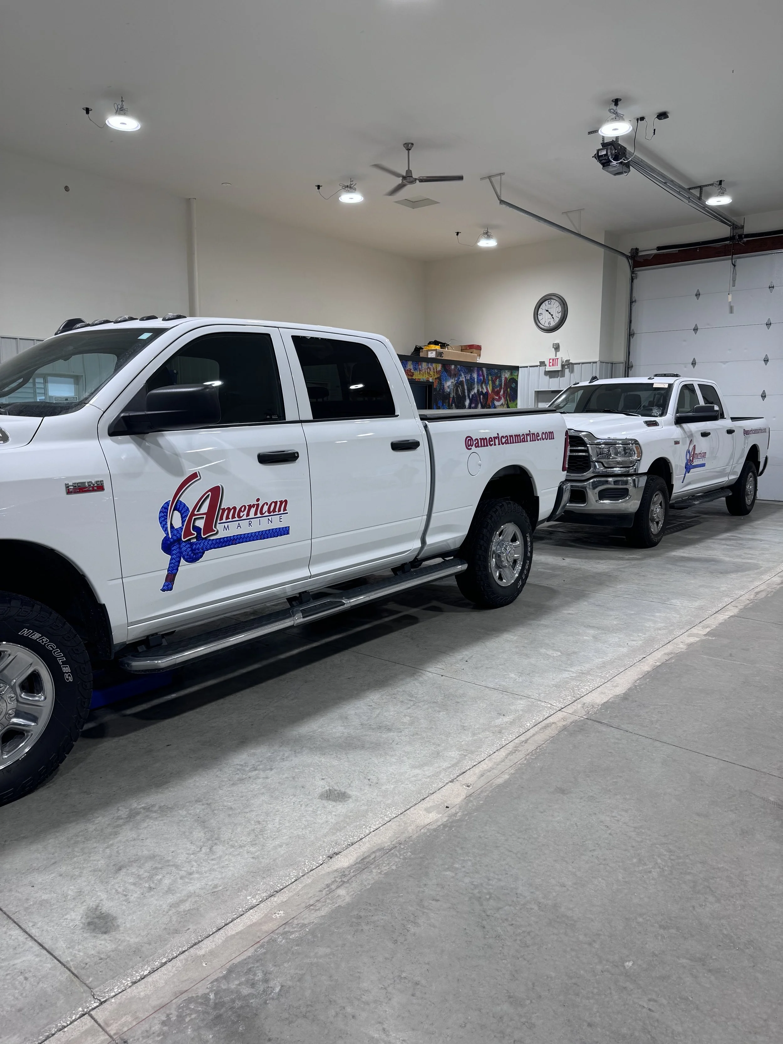 Two white pickup trucks with 'American Marine' logos and website URL parked inside a garage.