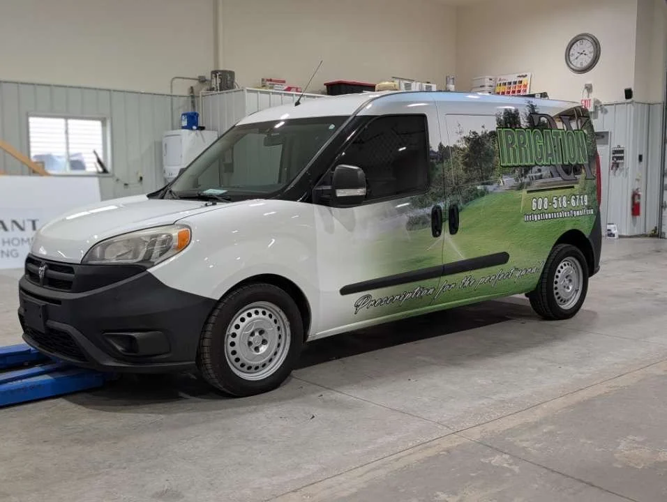 A white service van with green and black branding for irrigation services, parked indoors in a warehouse or garage, with a phone number and slogan visible on the side.