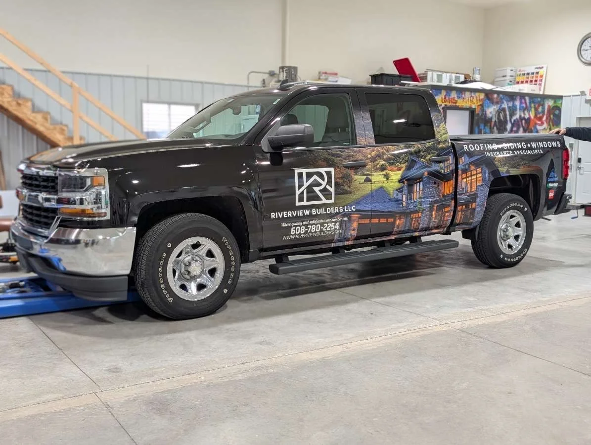 Black pickup truck with a dealership advertisement for Riverview Builders LLC, featuring a house and scenic landscape illustration, parked inside a garage or workshop.