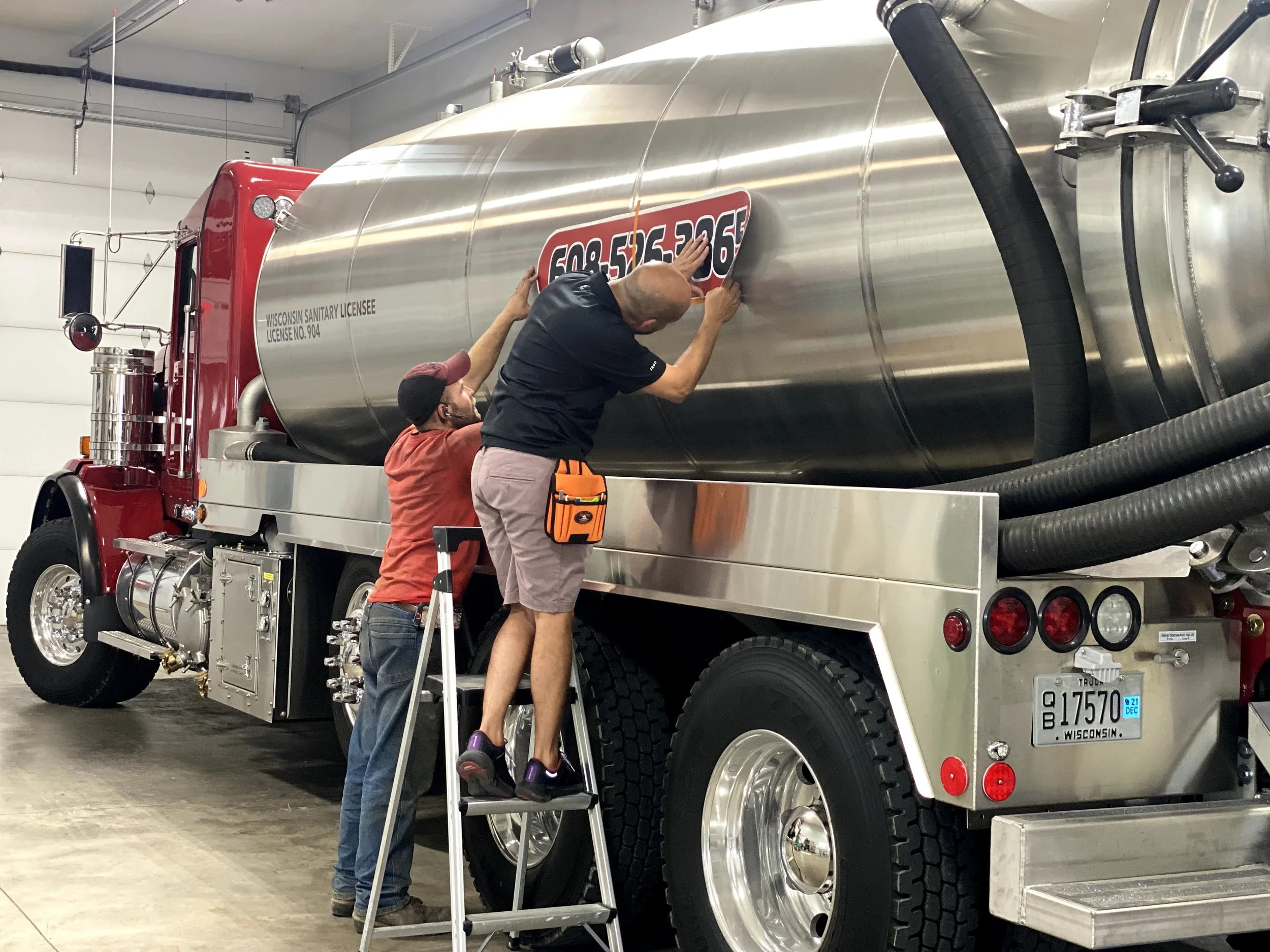 Two men are attaching a sign to a large, shiny tanker truck inside a garage or warehouse. One man is standing on a ladder, and the other is reaching up from the ground. The truck is red and silver with a Wisconsin license plate.