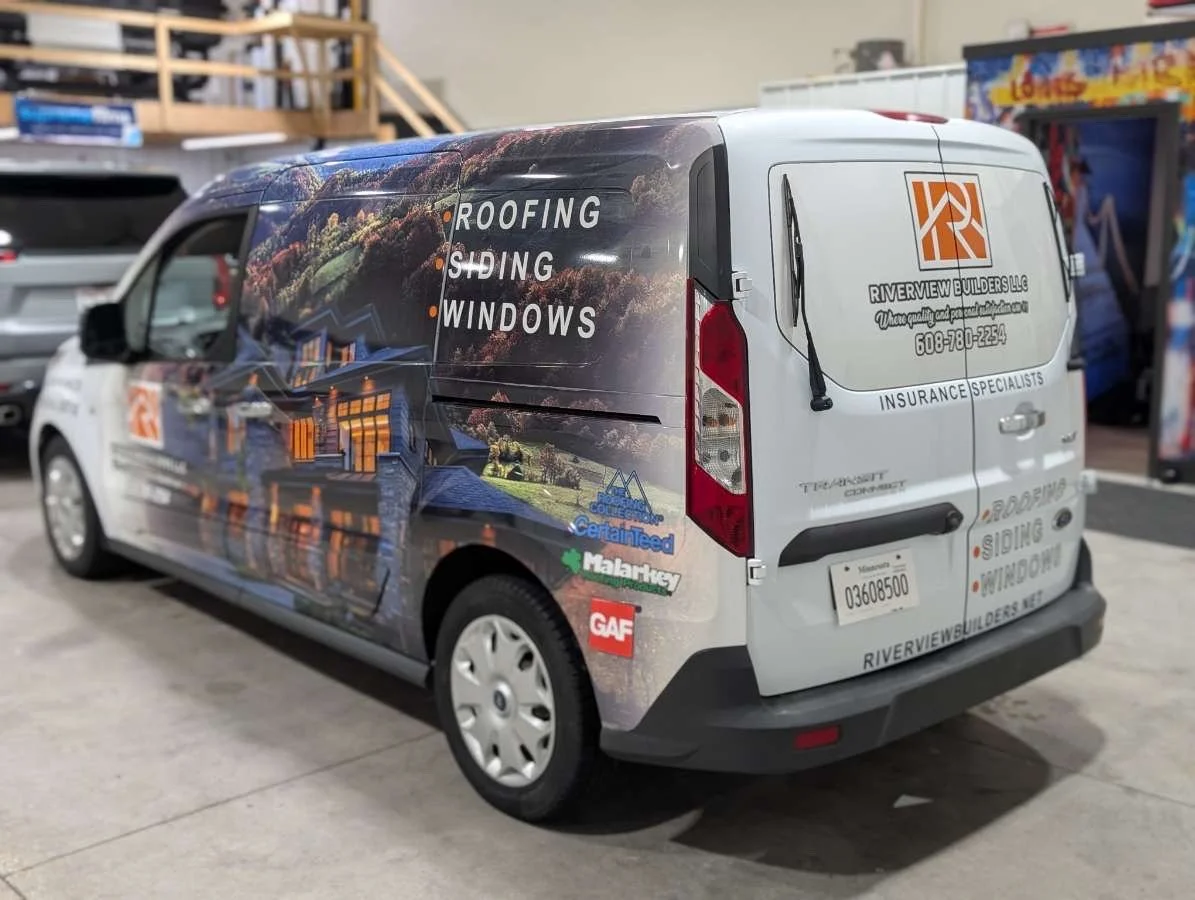 Commercial van with branding for Riverview Builders LLC, showing services like roofing, siding, and windows, parked indoors, with a large graphic of houses and trees on the vehicle's side.