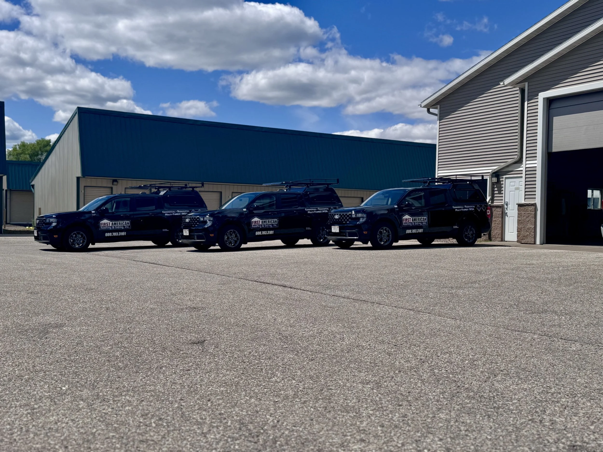 Three black company vehicles with First American Roofing signs parked outside a garage on a sunny day with partly cloudy sky.
