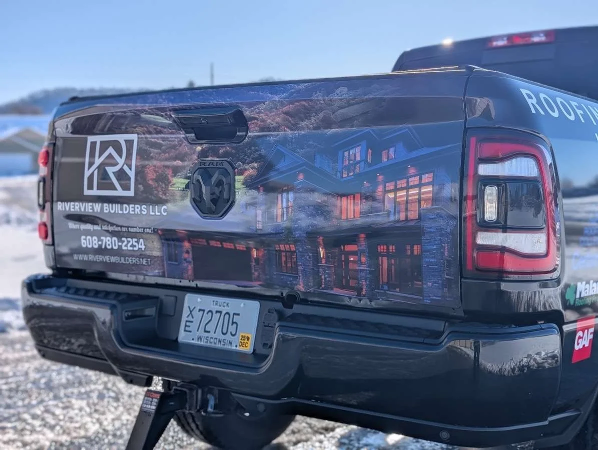 Truck with a vinyl wrap advertisement for Riverview Builders LLC featuring a house with lights on outside, a Wisconsin license plate, and a partially visible roof repair advertisement on the back of the vehicle.