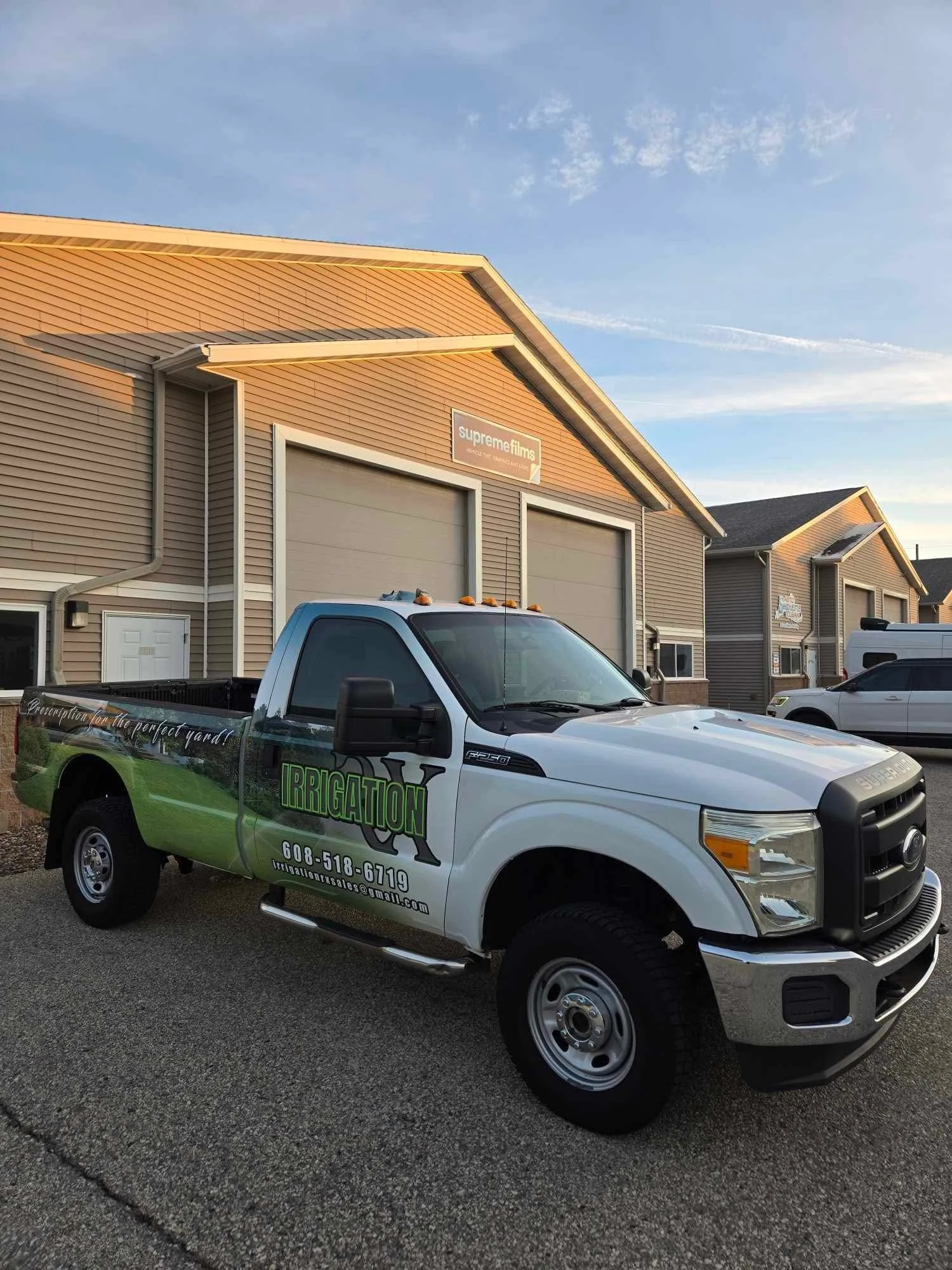 A white pickup truck with green and black lettering advertising irrigation services is parked in front of a beige industrial building with a sign that reads 'supreme films'.