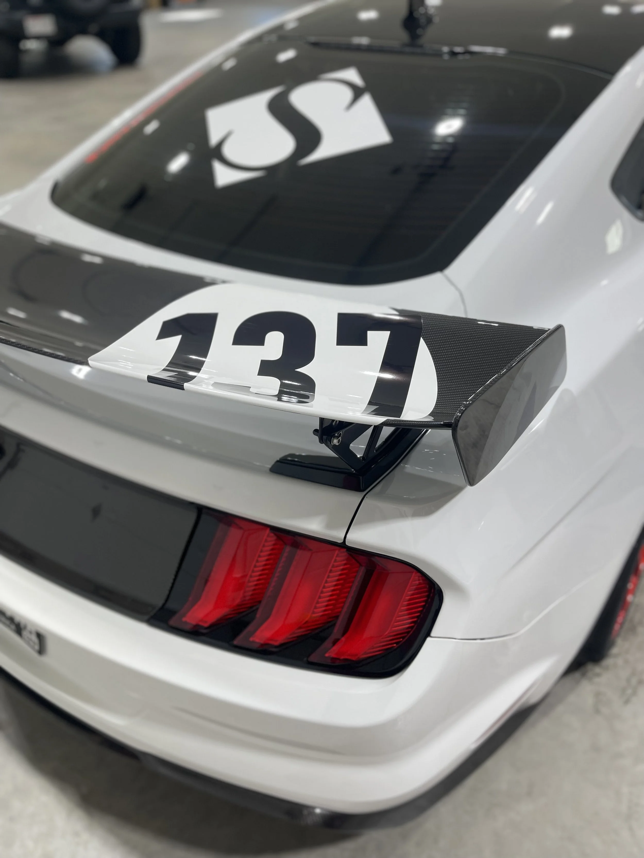 A white sports car with a racing spoiler featuring the number 137, parked indoors alongside other vehicles.