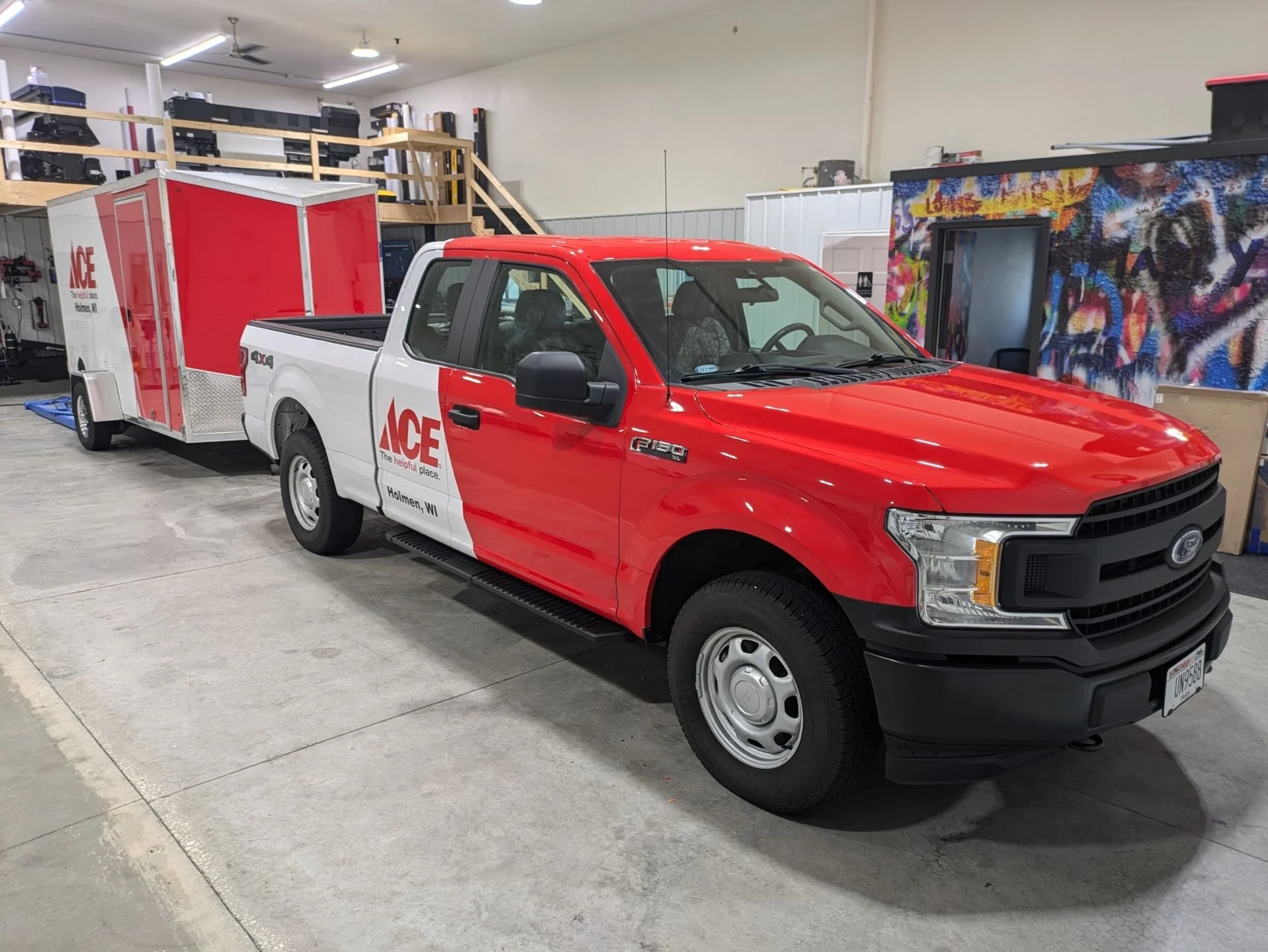 Red and white Ford F-150 pickup truck with a trailer attached, both branded with ACE Hardware logo, parked inside a warehouse.