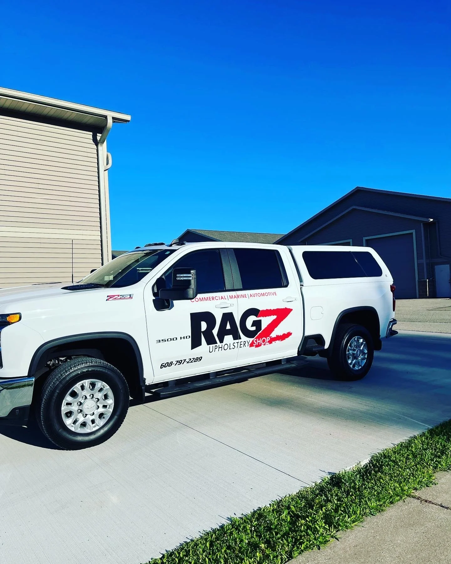White pickup truck with RAG Z Upholstery Shop advertisement parked on pavement in front of houses under a blue sky.