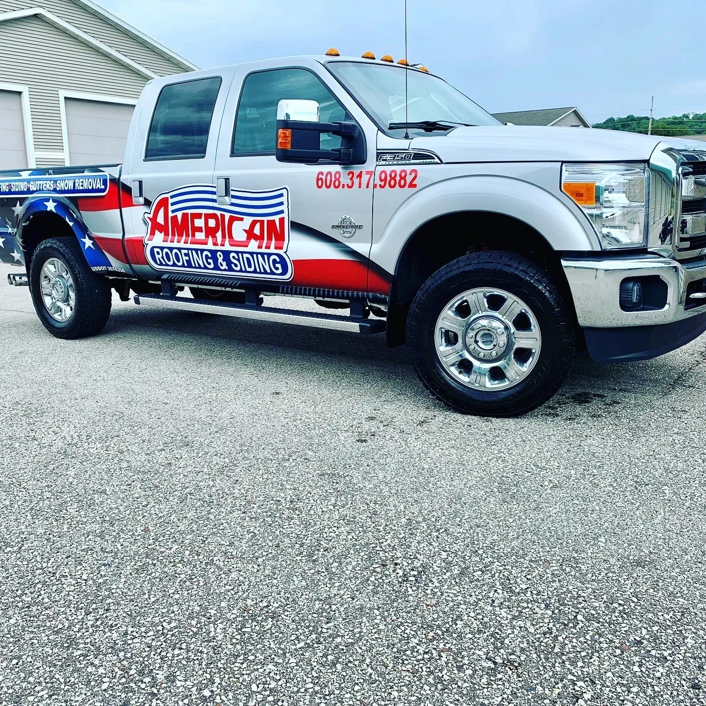 A white pickup truck with American flag-themed advertising for roofing and siding, parked outdoors on a gravel surface near a house, with the company name, phone number, and logo displayed.