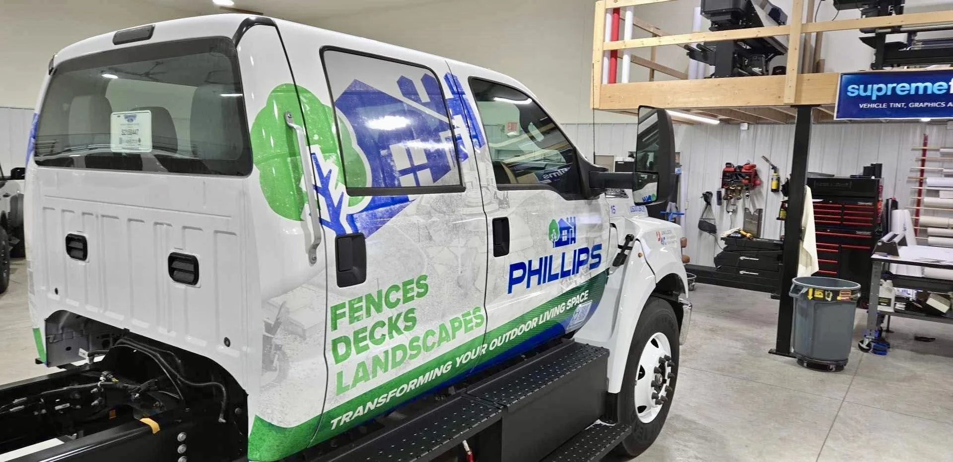 A white service vehicle with graphic decals for Phillips, promoting fences, decks, landscapes, and outdoor living space, inside a workshop with tools, shelving, and equipment.