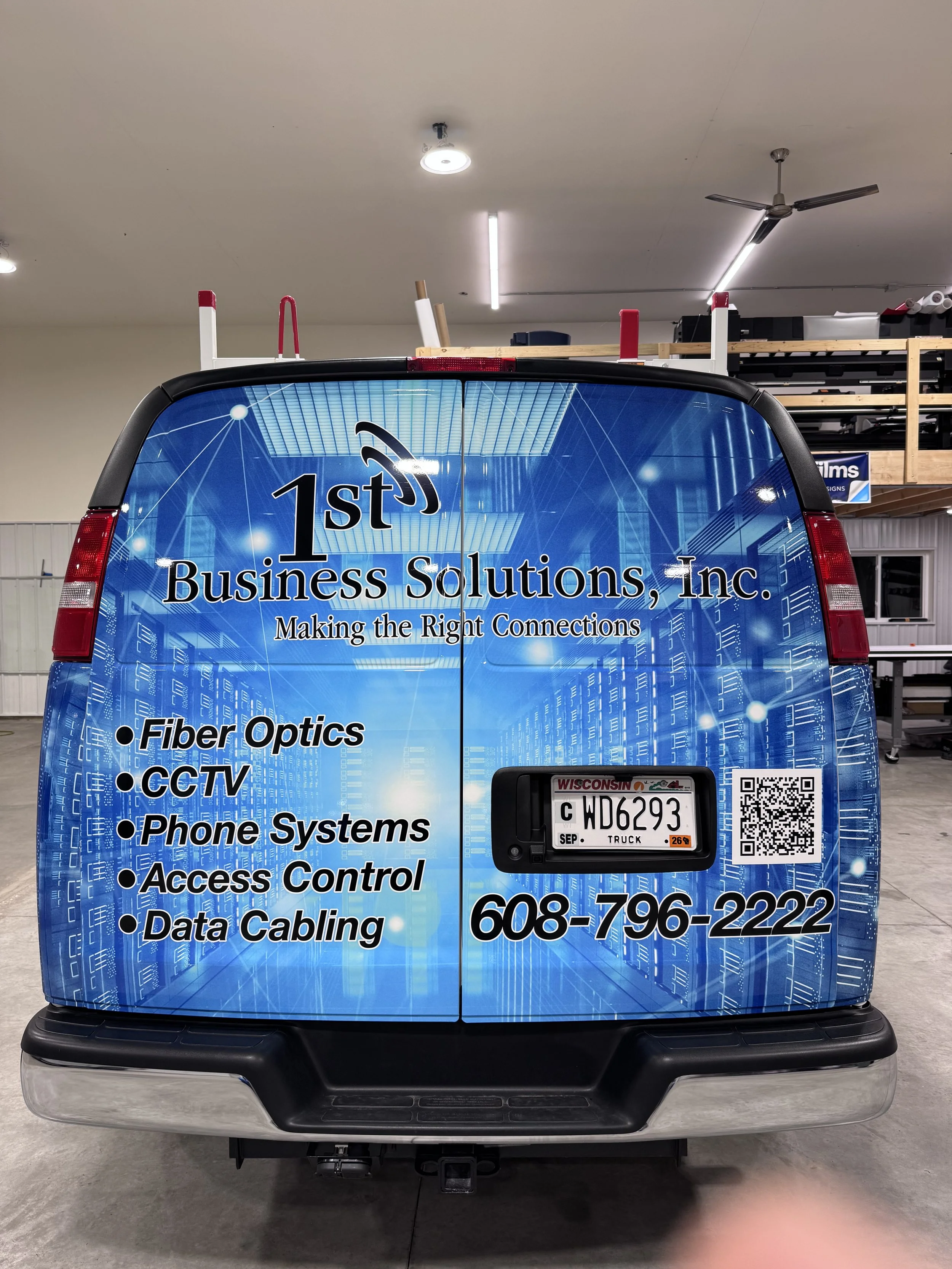 The back of a business van with a blue digital-themed wrap displaying the company's name, contact information, and services including fiber optics, CCTV, phone systems, access control, and data cabling.