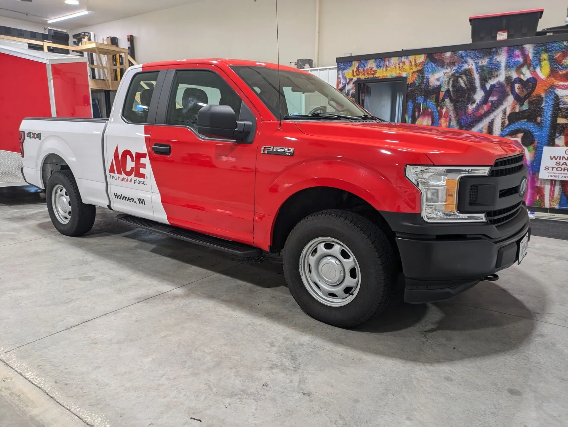A red and white pickup truck with ACE logo parked indoors. The truck has a black grille, black side mirrors, and the text 'Holmen, WI' on the door.